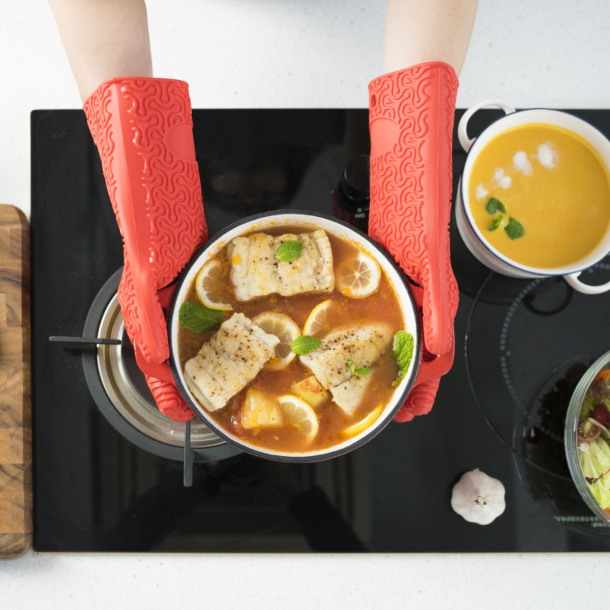 Rote Ofenhandschuhe aus Silikon beim Halten eines Topfs mit Suppe auf Herdplatte.