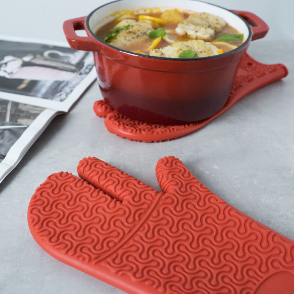 Roter Ofenhandschuh aus Silikon mit Strukturmuster auf grauem Untergrund neben rotem Kochtopf mit Eintopf.