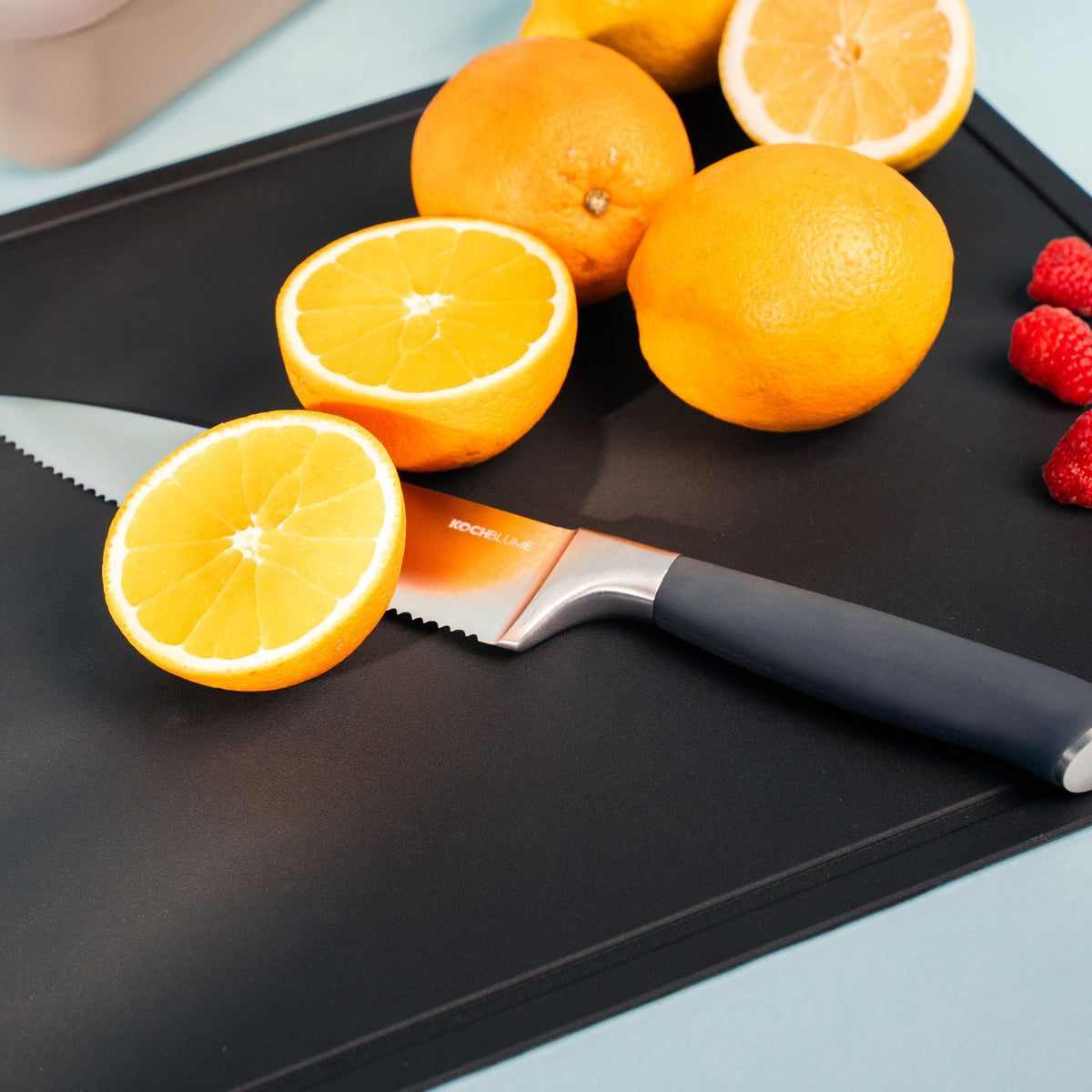 Schwarze rechteckige Schneidunterlage mit geschwungenem Messer, darauf Orangen und Himbeeren.