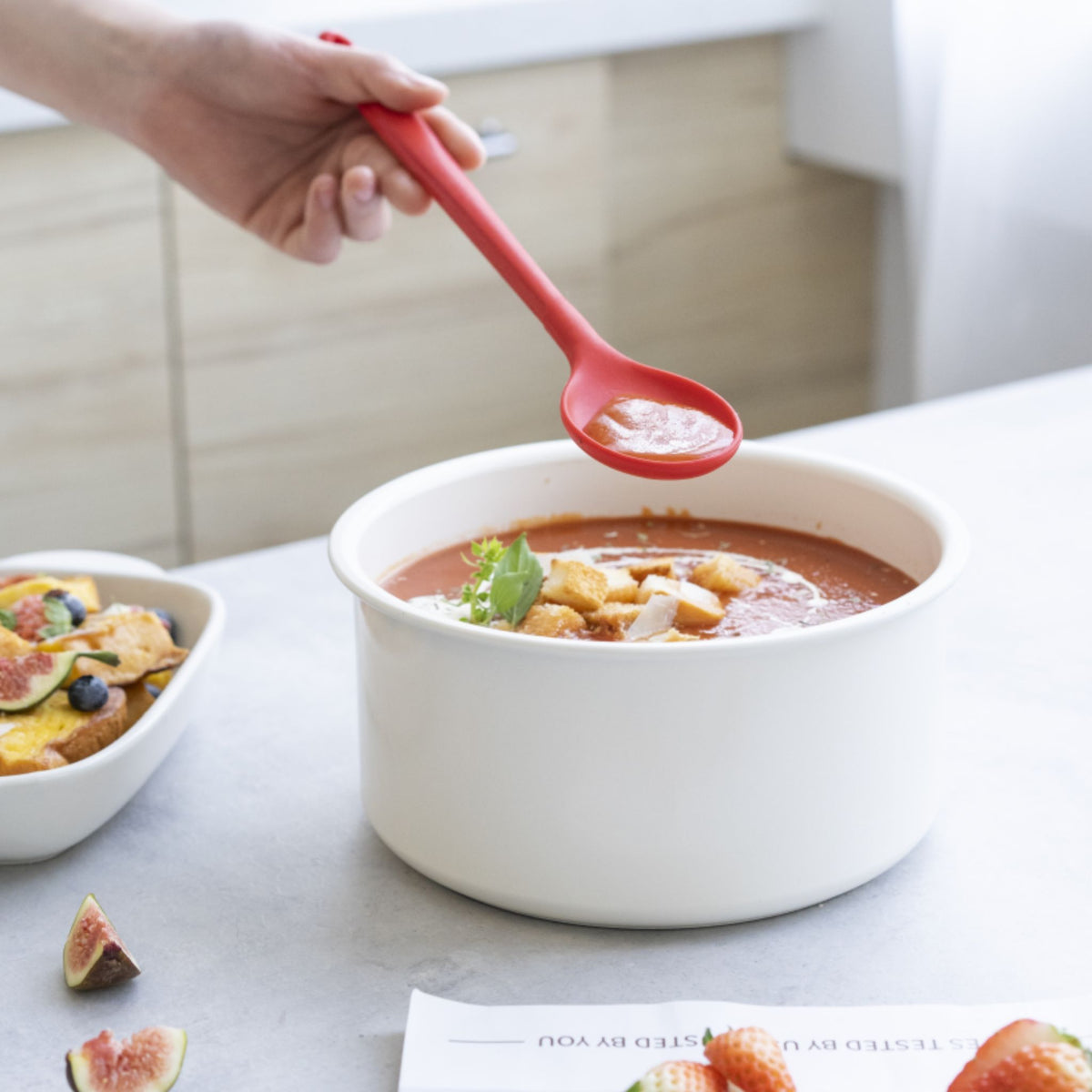 Roter Kochlöffel aus Silikon über weißem Kochtopf mit Suppe und Einlage.