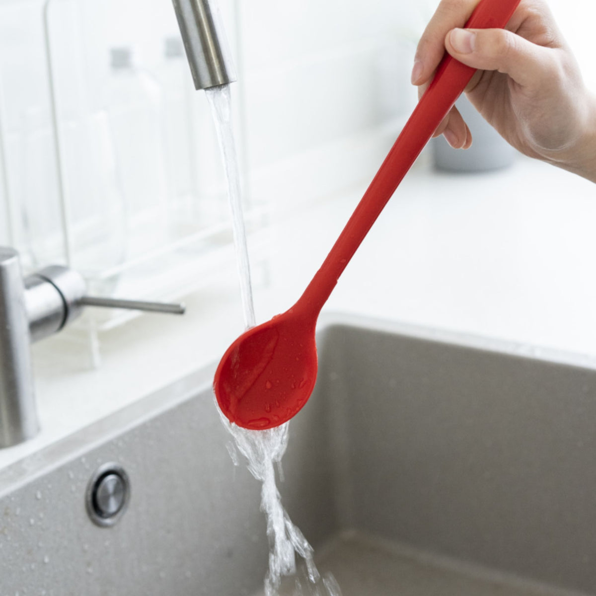Roter Kochlöffel aus Silikon mit langem Griff unter fließendem Wasser über Spüle gehalten.