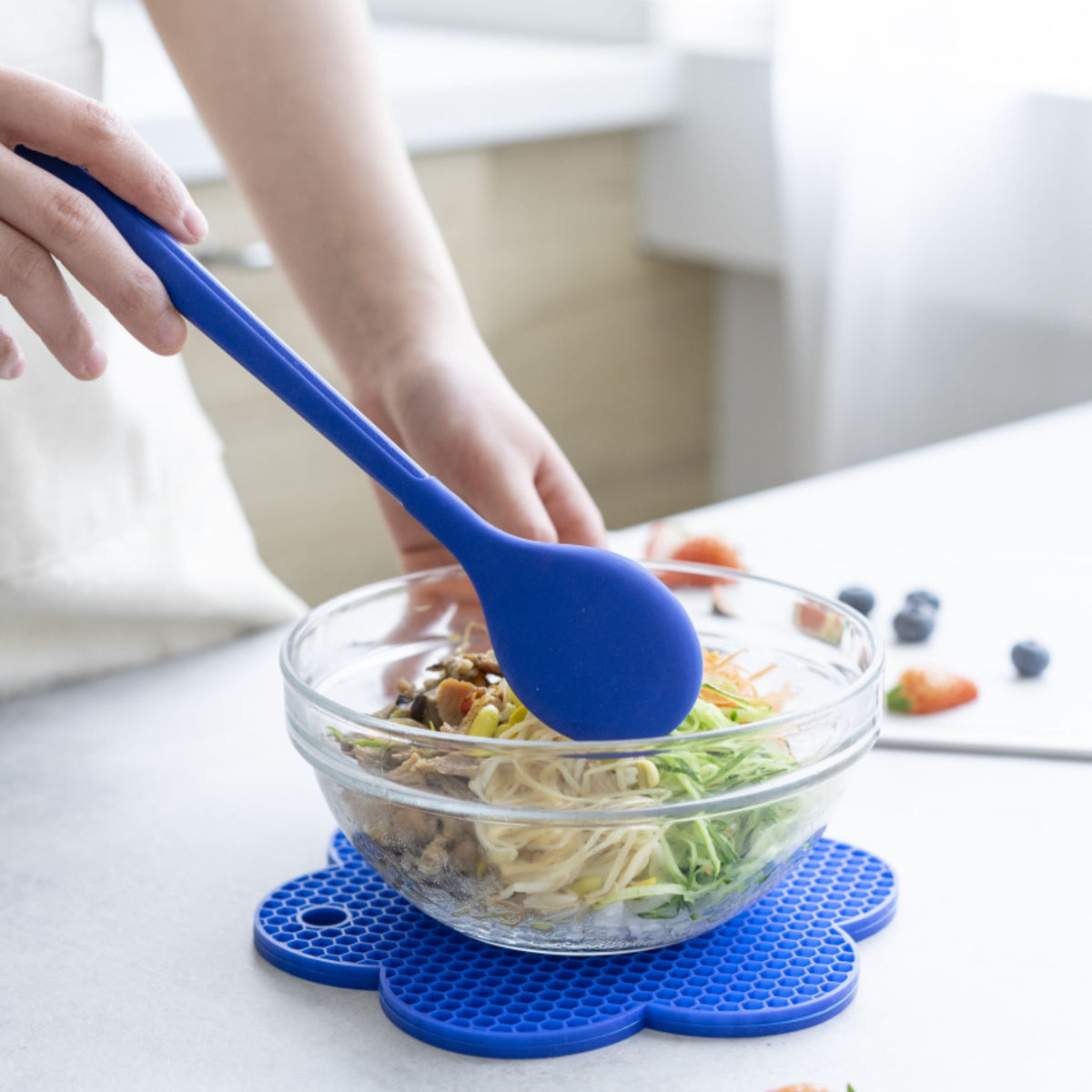 Blauer Kochlöffel aus Silikon rührt Salat in glasschüssel auf blauer Unterlage.