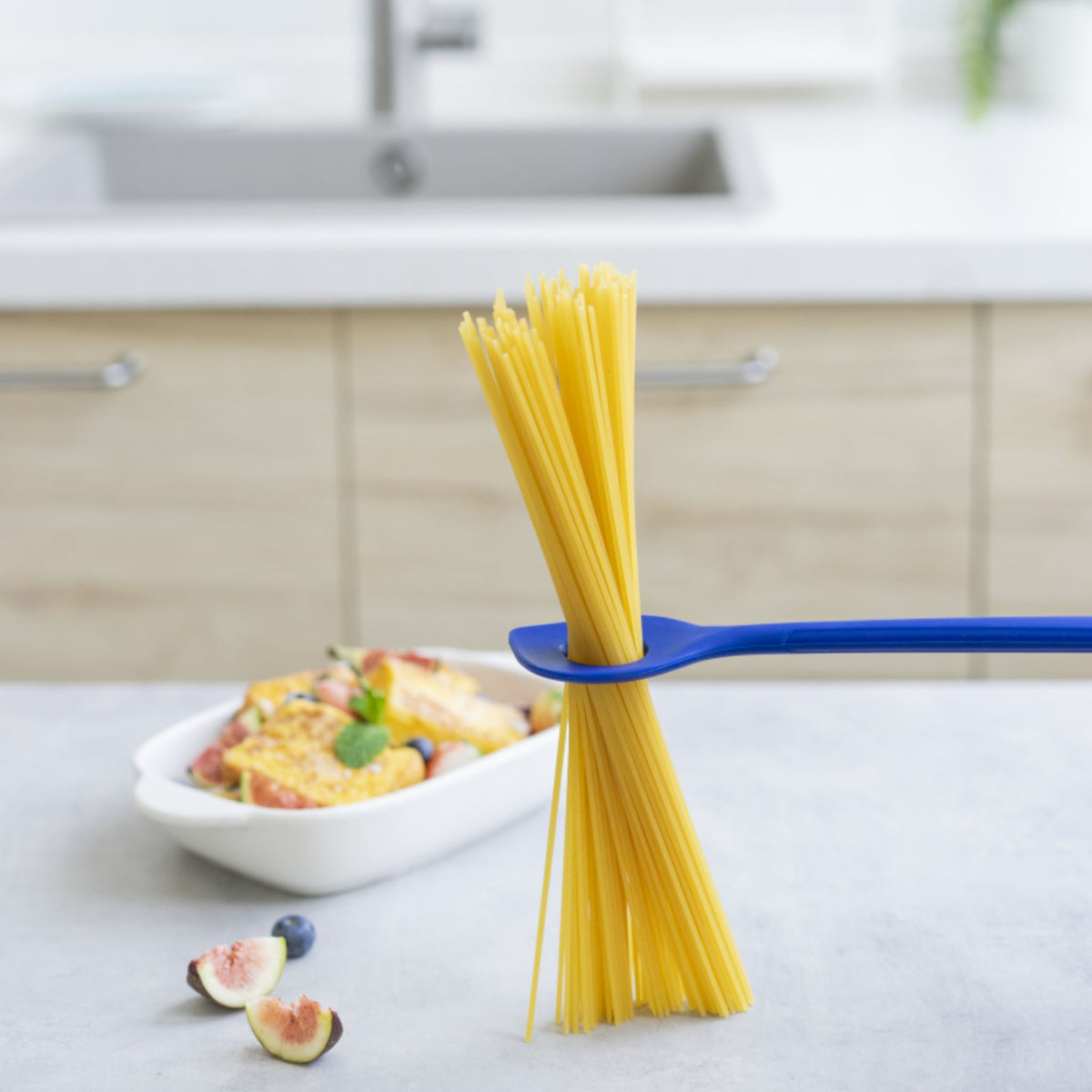 Blauer Backlöffel hält ungekochte Spaghetti mit weißer Auflaufform im Hintergrund.