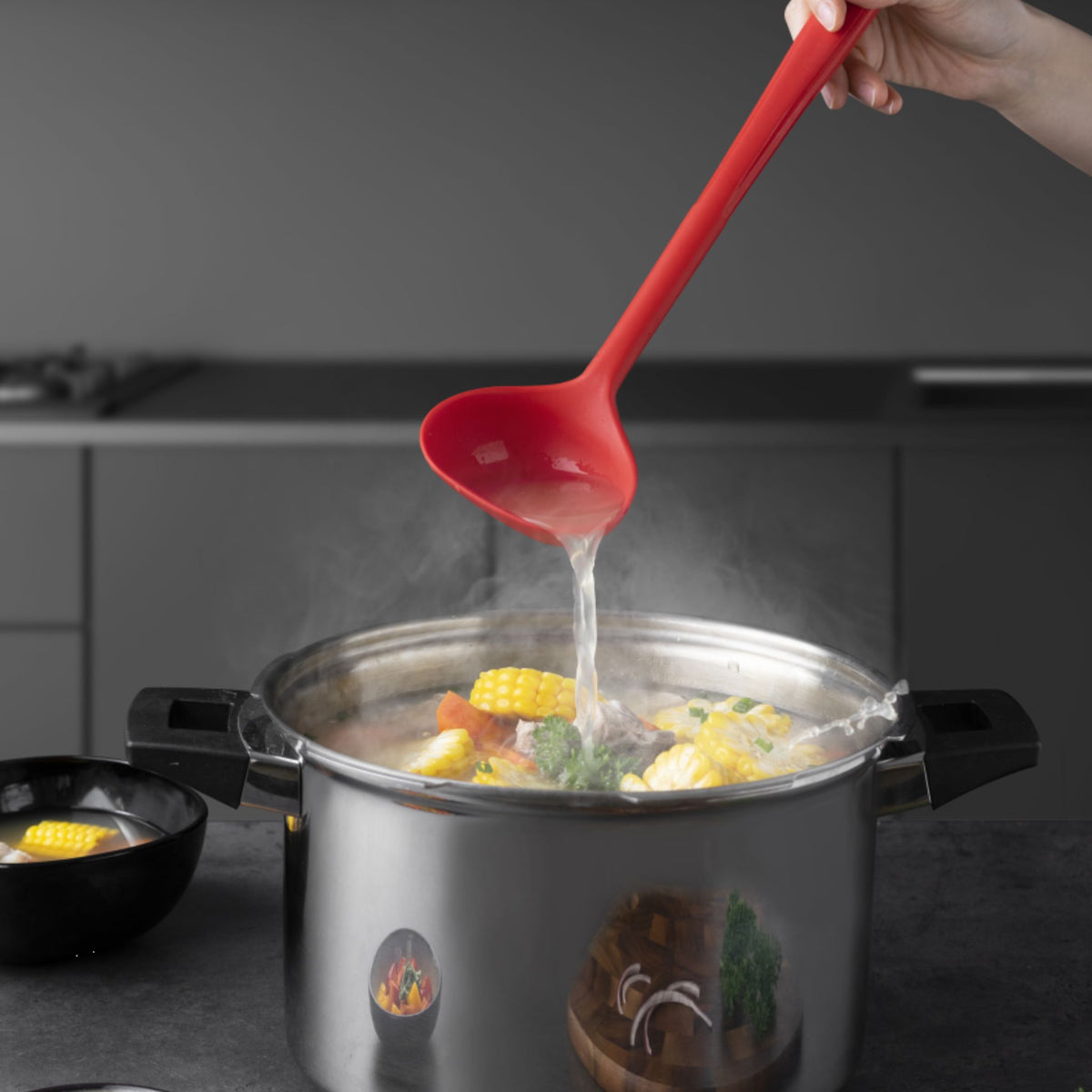 Rote Schöpfkelle aus Kunststoff gießt Suppe aus dampfendem Topf mit Gemüse.