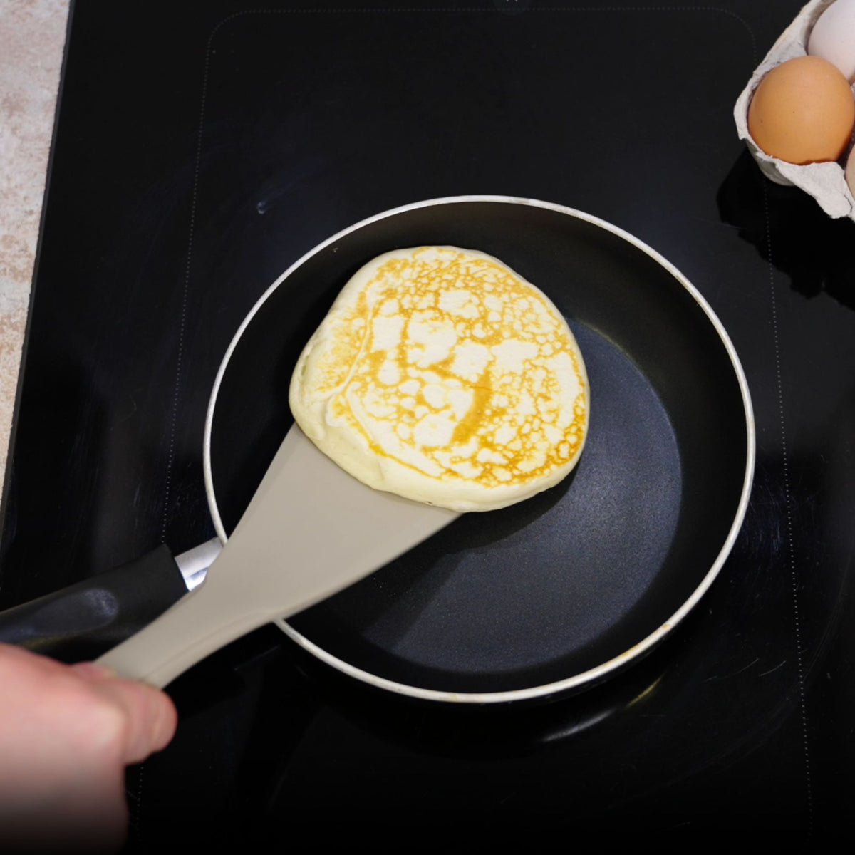 Anthrazitfarbener Pfannenwender mit breiter Klinge wendet einen Pfannkuchen in einer schwarzen Pfanne.