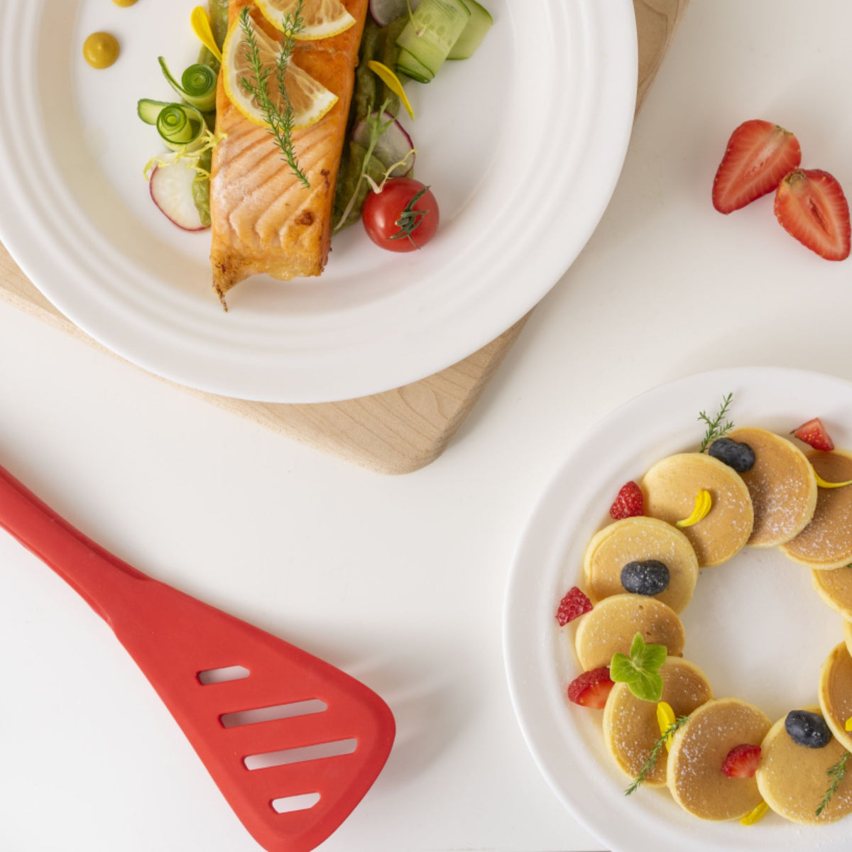 Roter Pfannenwender mit Schlitz neben Teller mit Lachs und Pancake-Teller mit Beeren und Minze.