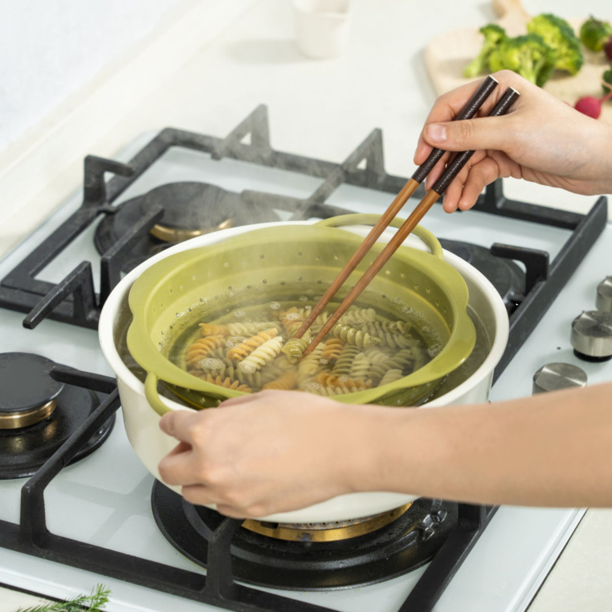 Grünes Faltsieb aus Silikon mit Henkel beim Abseihen von Pasta auf Herd.