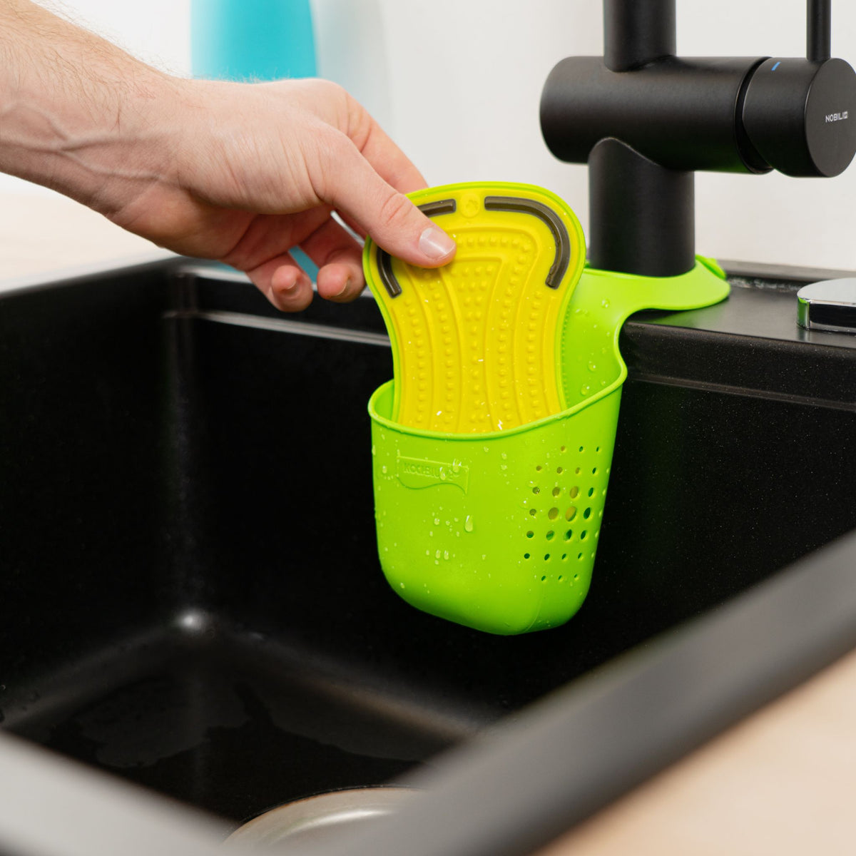 Anthrazitfarbener Schwammhalter aus Kunststoff am Wasserhahn montiert mit eingesetztem gelben Schwamm.