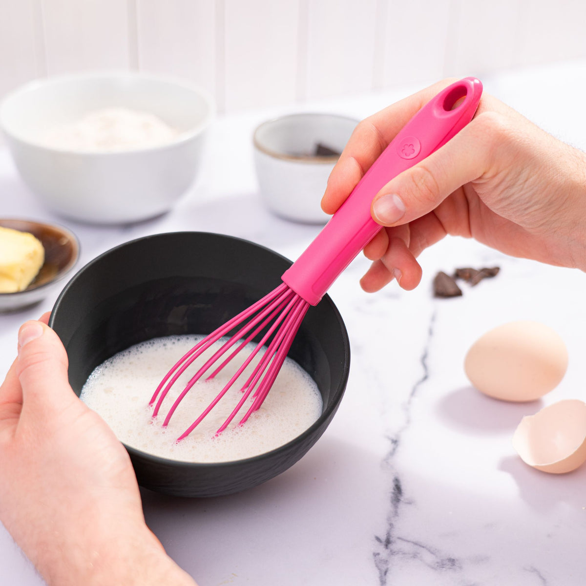 Schneebesen in Pink mit Silikongriff und Drahtschlaufen in schwarzer Schüssel mit weißer Flüssigkeit.