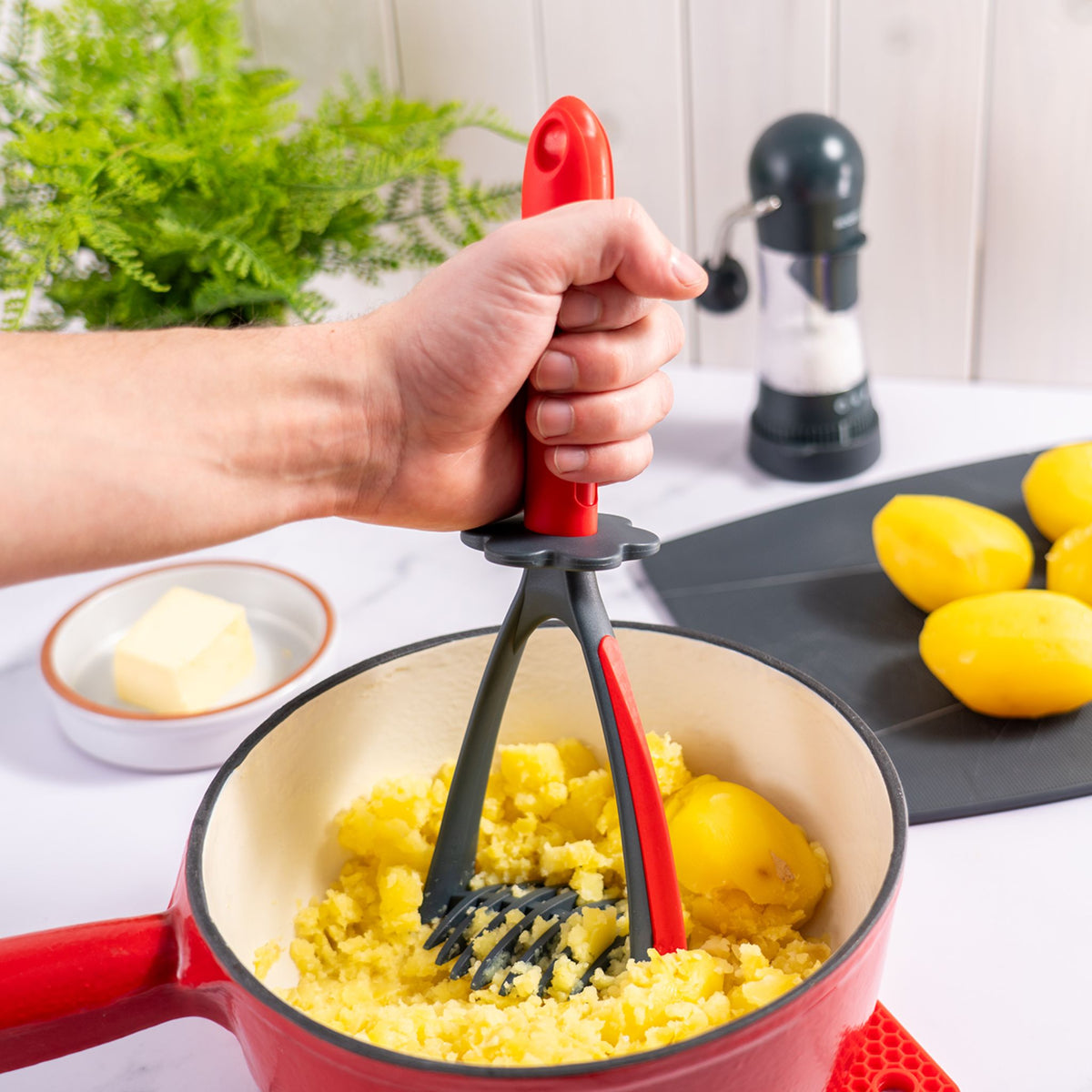 Kartoffelstampfer mit rotem Griff zerdrückt Kartoffeln in weißem Topf mit rotem Rand.