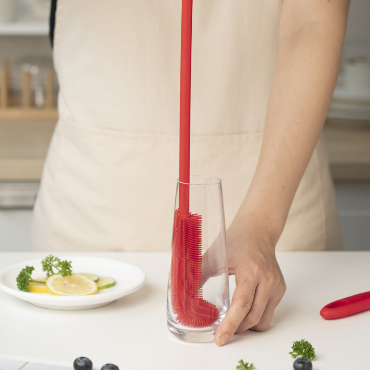Rote dünne Flaschenbürste mit langem Stiel in klarem Glasbecher auf Küchentheke.