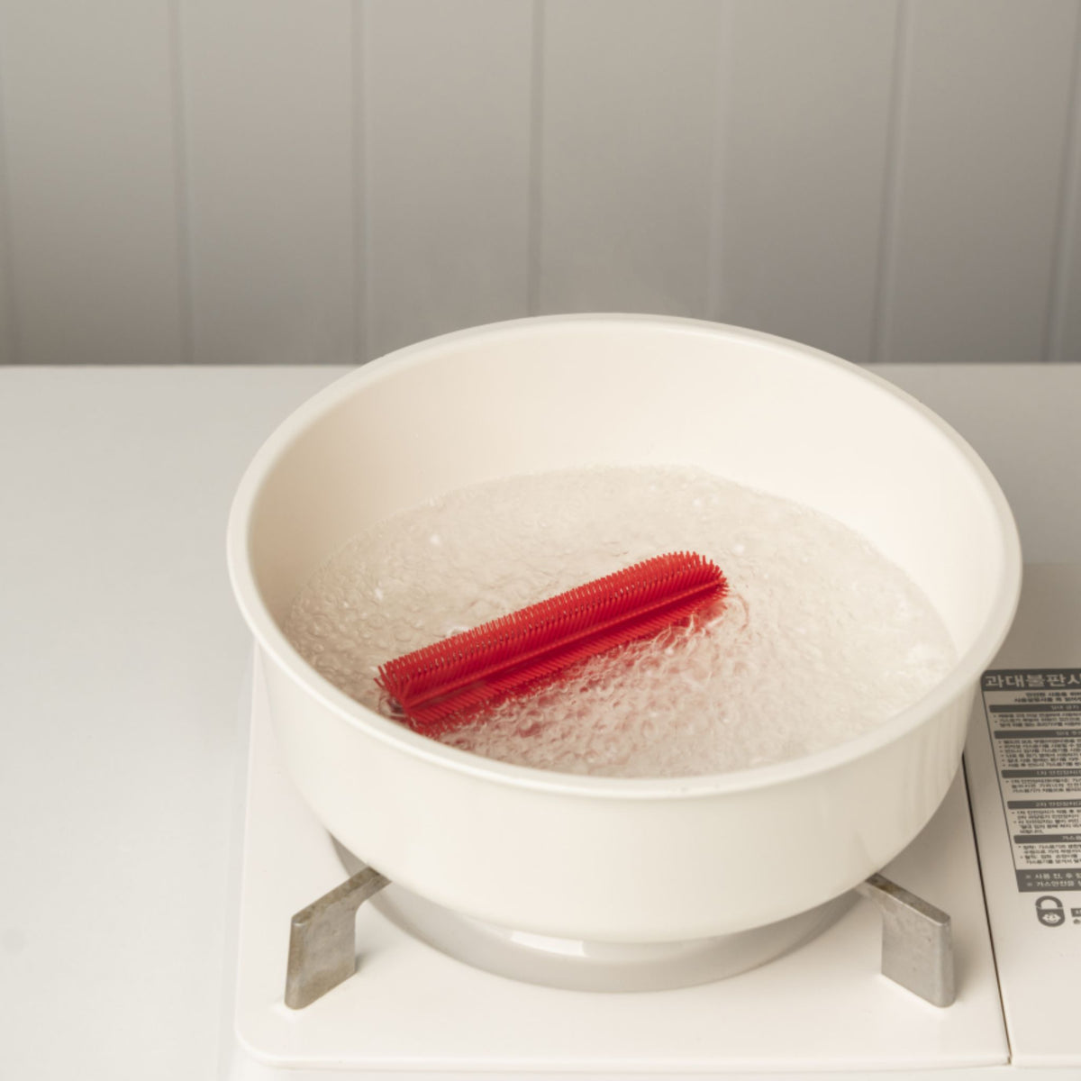 Rote dünne Flaschenbürste in weißem Topf mit sprudelndem Wasser auf Herd.