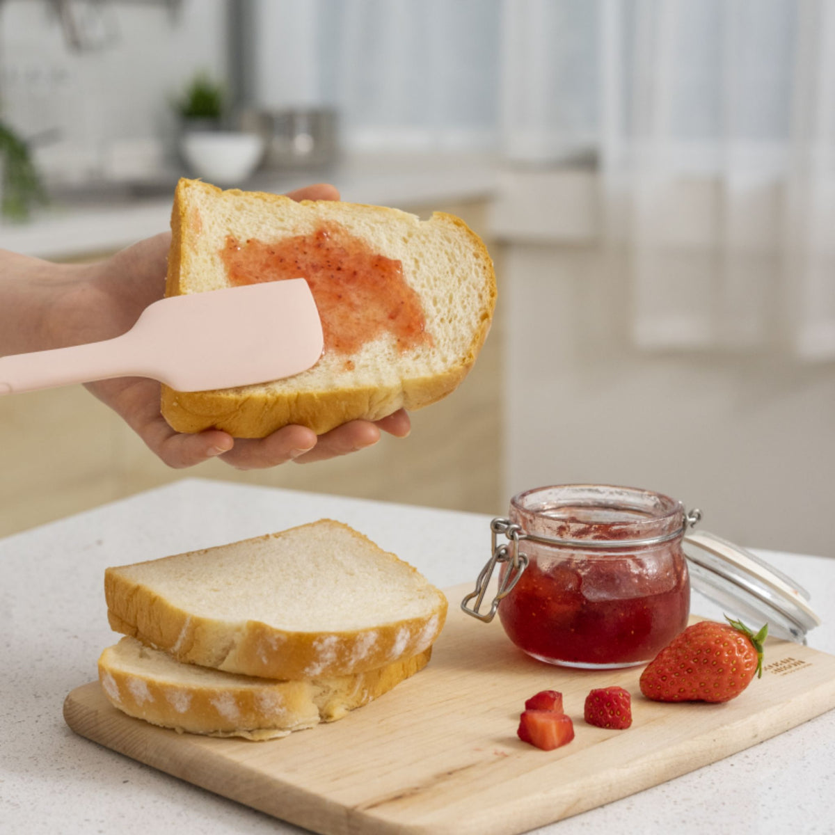 Altrosa Teigschaber mit flexiblem Kopf streicht Erdbeermarmelade auf Weißbrotscheibe.