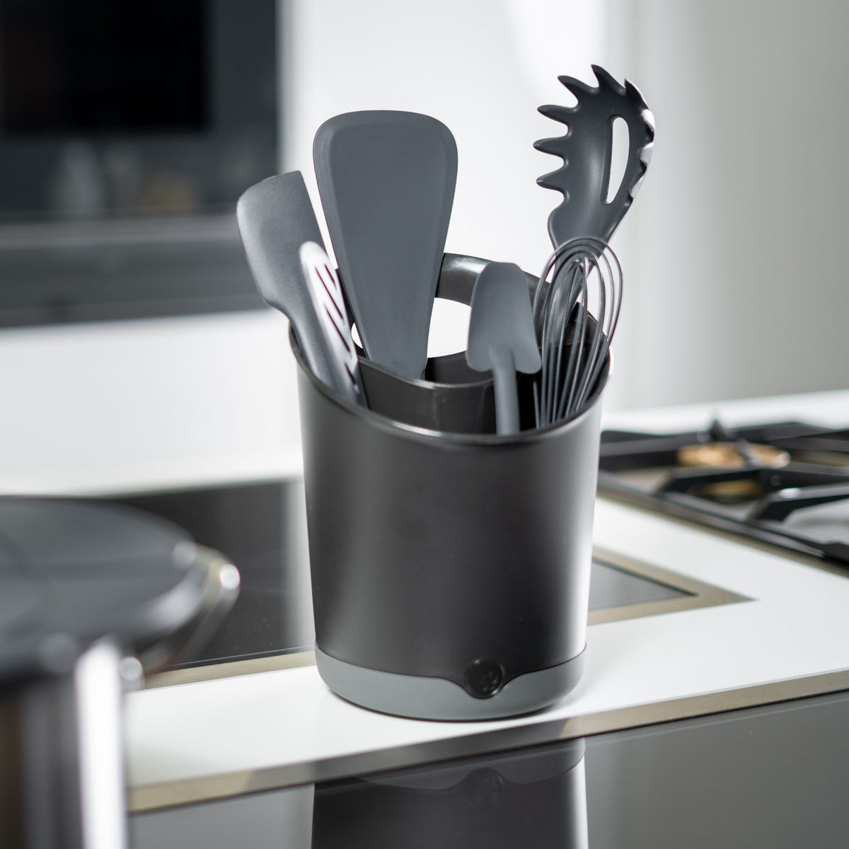 Schwarzer Utensilienbehälter mit grauen Küchenhelfern aus Silikon und Metall auf weißer Arbeitsplatte.