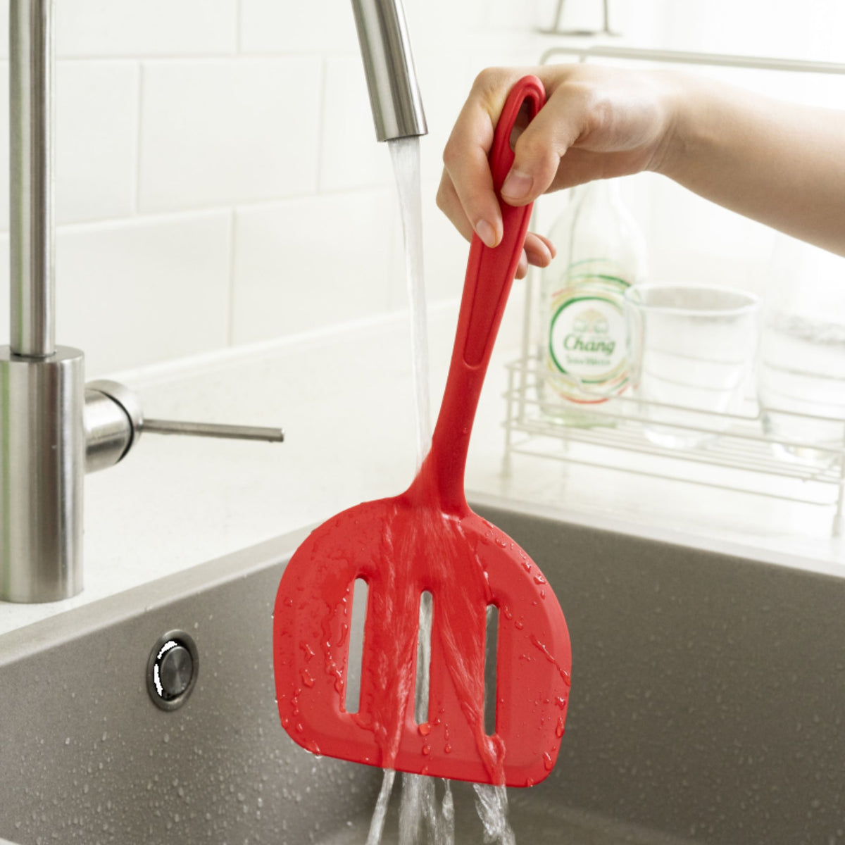 Roter Fischwender aus Kunststoff mit langem Griff unter fließendem Wasser in der Spüle.