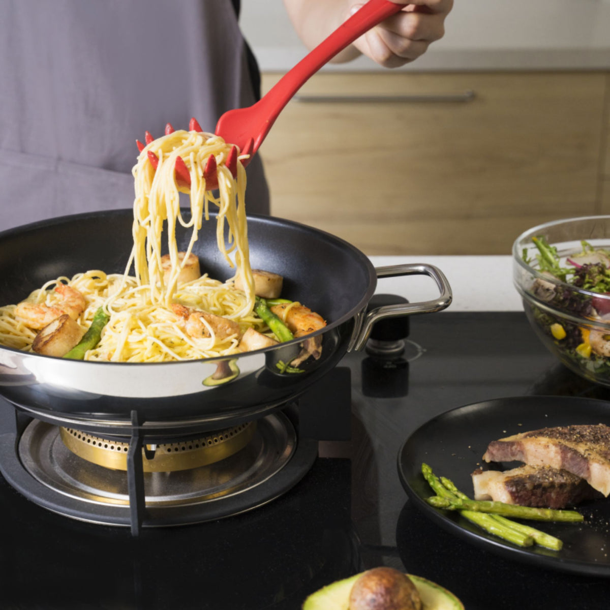 Roter Pastalöffel hebt Spaghetti mit Soße aus schwarzer Pfanne auf Herd.