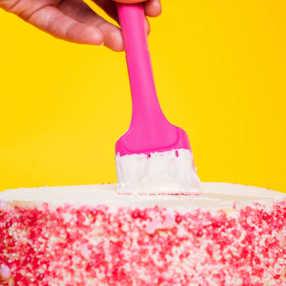 Backpinsel mit pinkem Griff und weißen Silikonborsten, benutzt auf weißem Kuchen mit roten Streuseln.