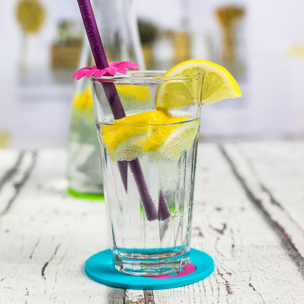 Glas mit Wasserglas, Zitronenscheiben und lila Strohhalm auf türkisem Untersetzer mit pinkem Rand.