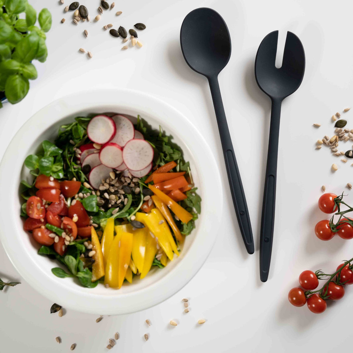Salatbesteck in Anthrazit neben bunter Salatschüssel mit frischem Gemüse und Saaten.