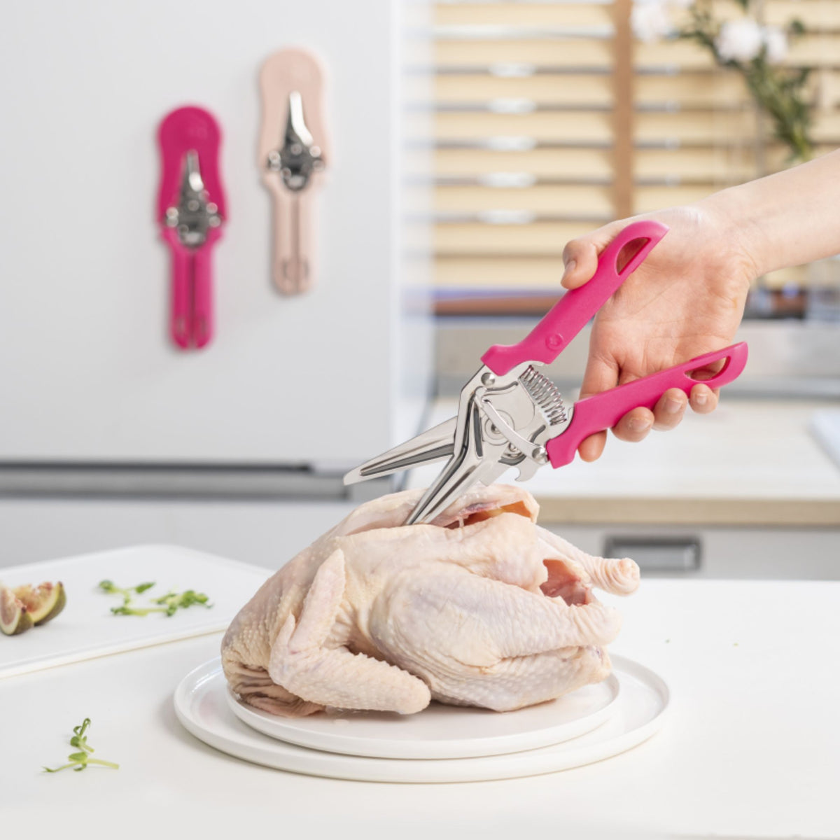Küchenschere mit pinken Griffen schneidet rohes Hähnchen auf weißem Teller.
