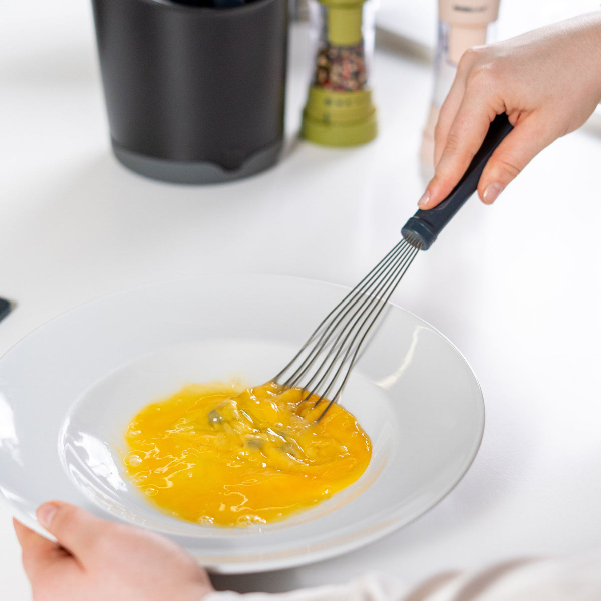 Anthrazitfarbener Tellerbesen aus Metall mit ergonomischem Griff, verwendet zum Schlagen von Eiern in weißem Teller.
