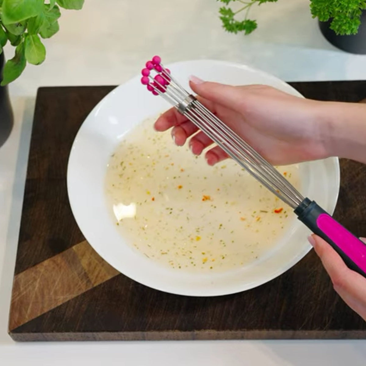Kugelschneebesen mit pinkem Griff und flexiblen Drahtenden über weißer Schüssel mit Flüssigkeit.