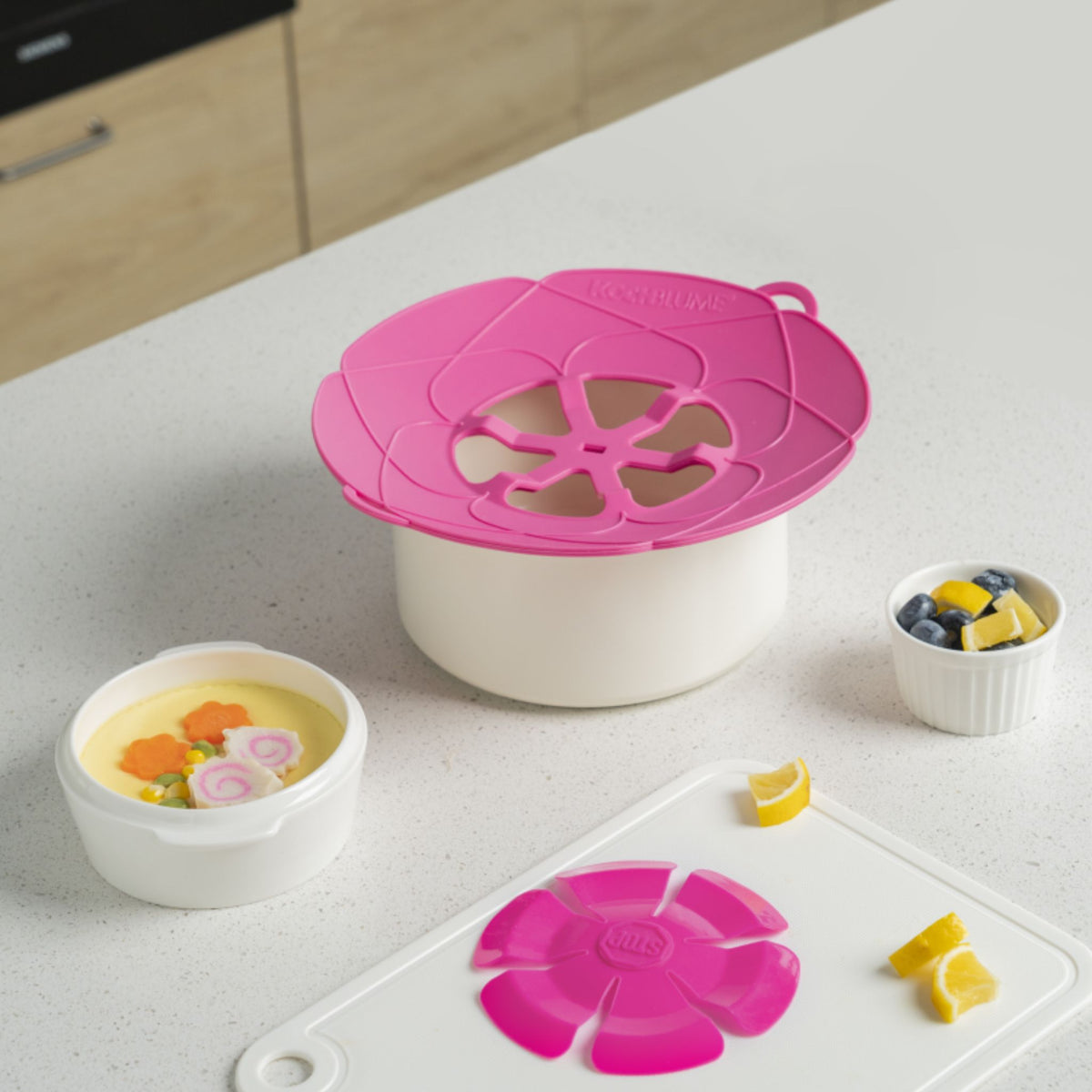 Runde Silikon-Kochblume in Pink auf weißem Topf mit Lebensmittel in Schalen auf Tisch.