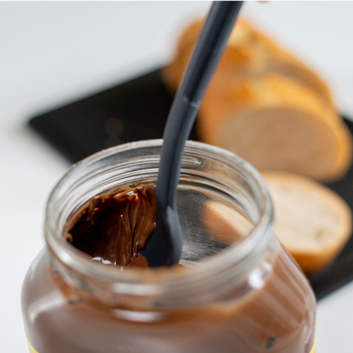 Anthrazitfarbener Restelöffel in Glas mit Schokoladencreme, unscharfe Brotscheiben im Hintergrund.
