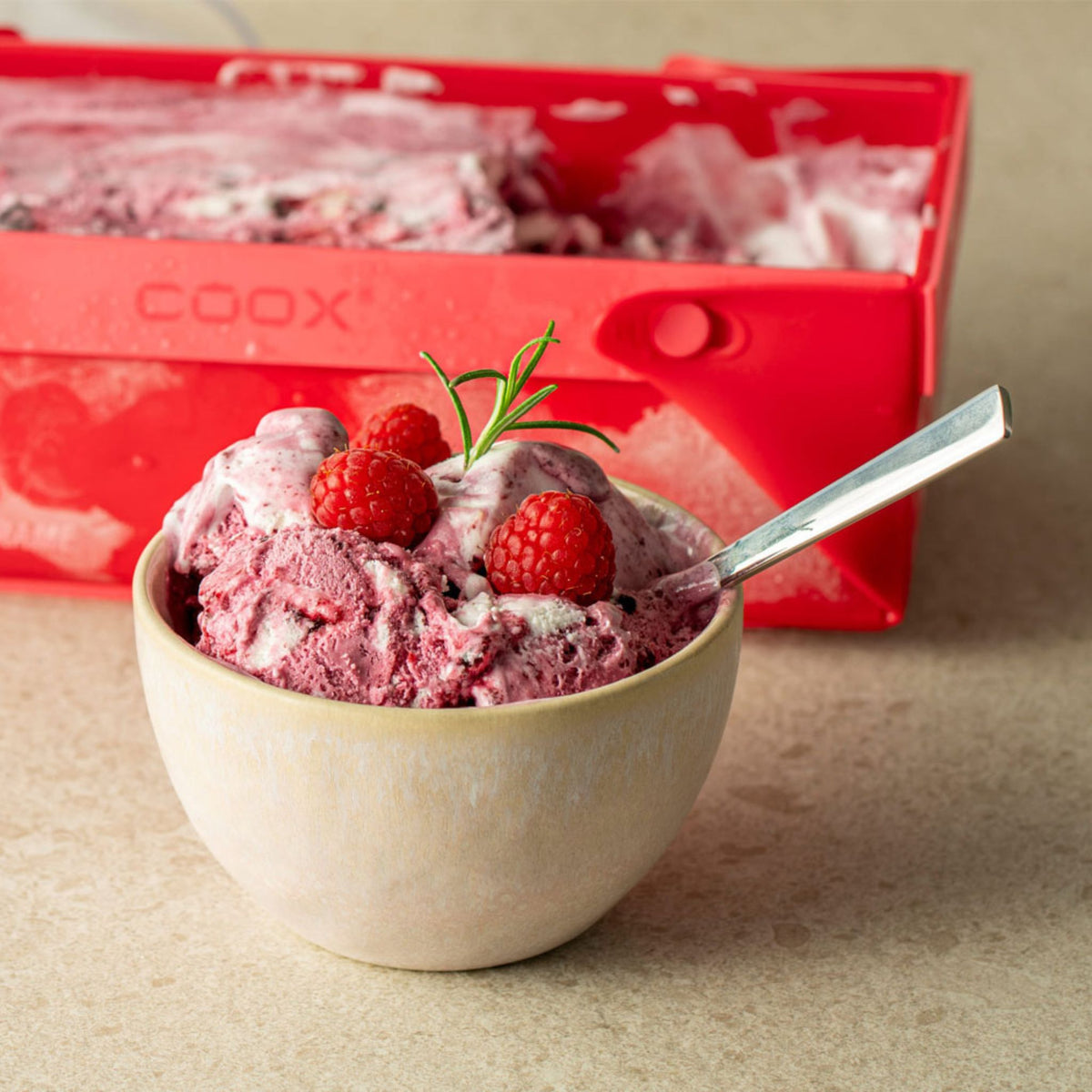 Beige Schale mit rosa Eis, garniert mit Himbeeren und Rosmarin, vor roter Verpackung.