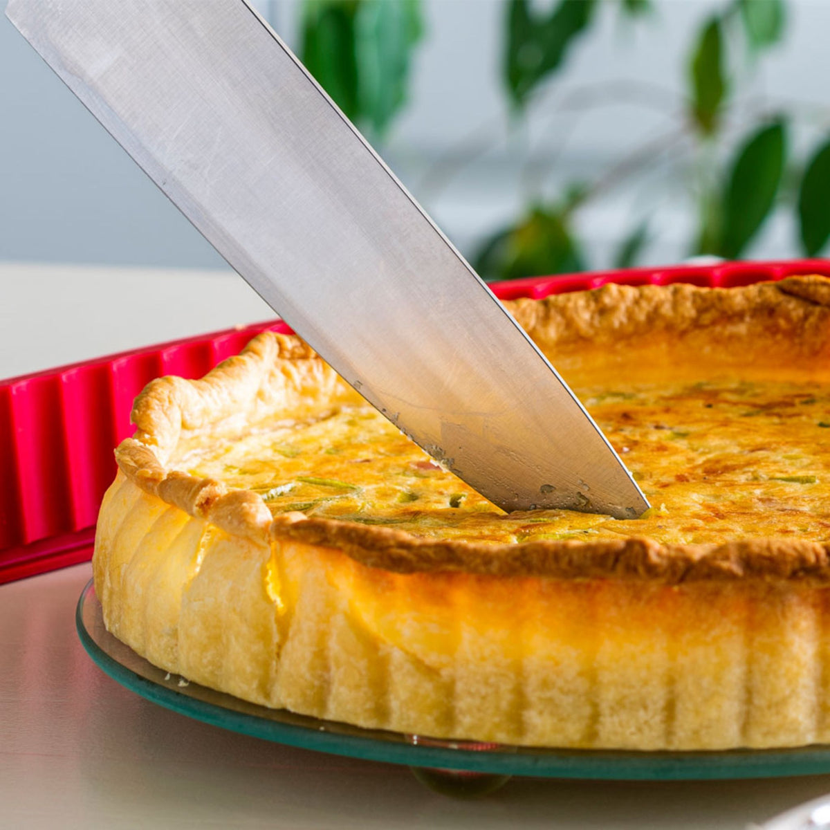 Rote Quicheform aus Metall mit gezacktem Rand und goldbraun gebackener Quiche beim Anschneiden.