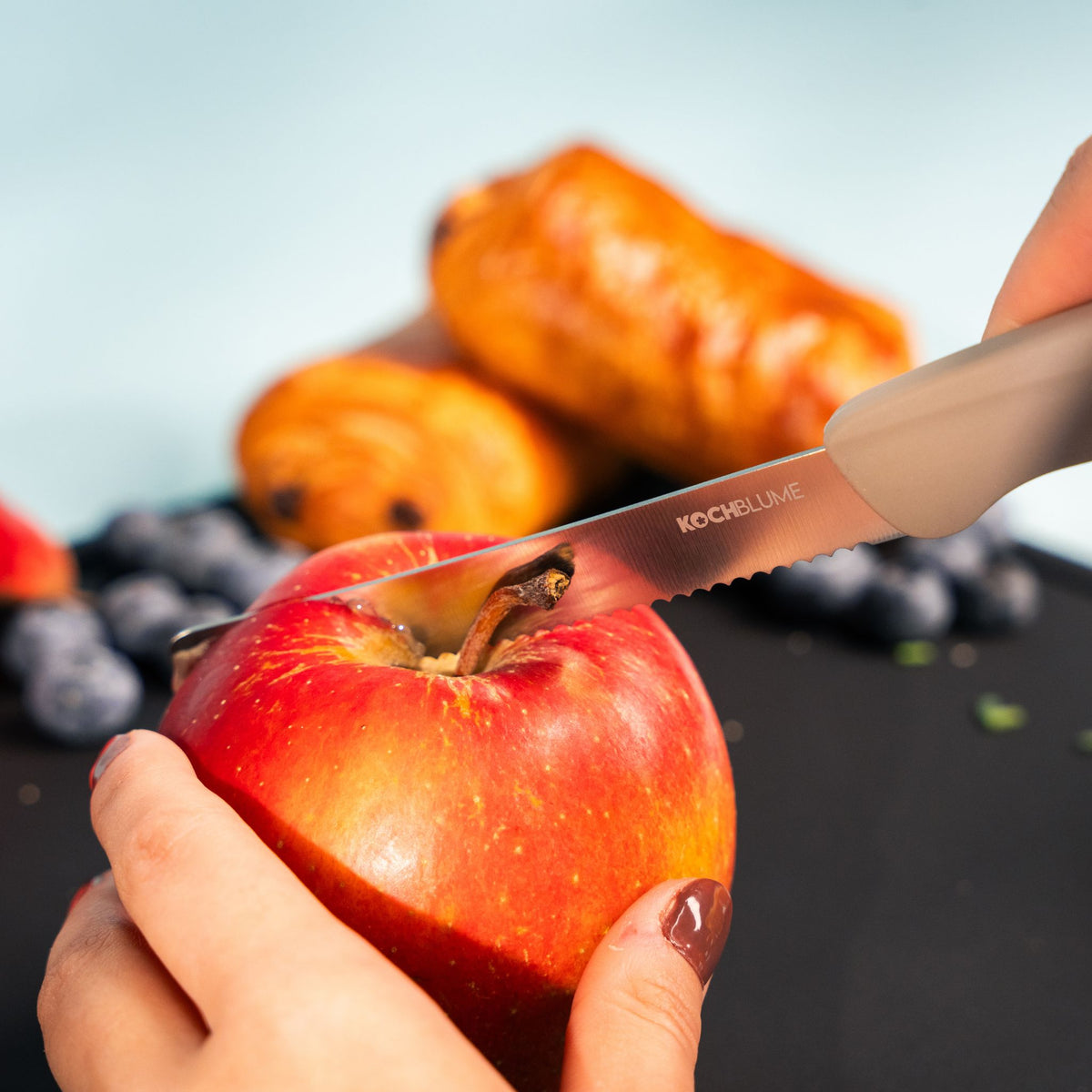 Frühstücksmesser mit gezahnter Klinge und taupefarbigem Griff schneidet roten Apfel.
