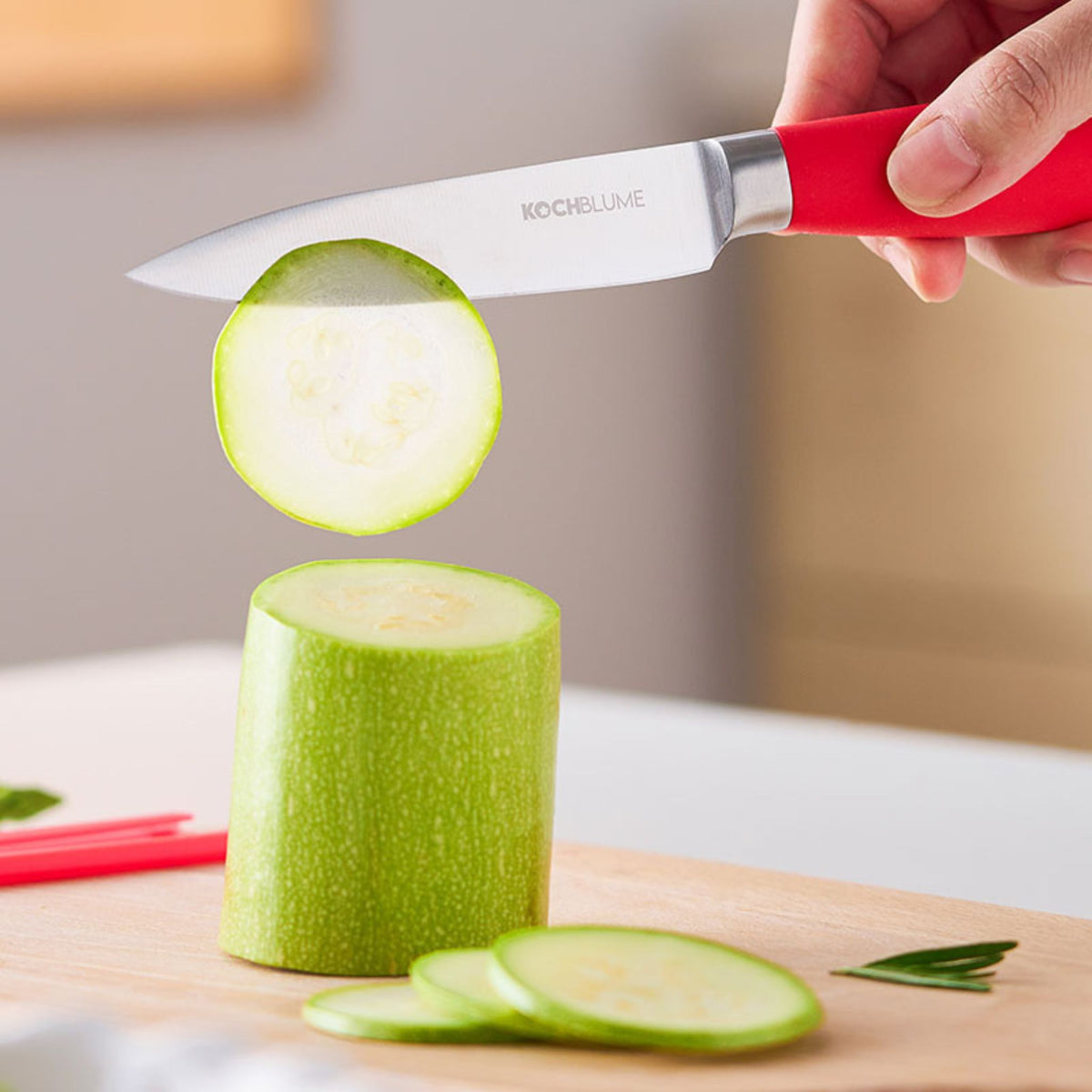 Gemüsemesser mit rotem Griff schneidet grüne Zucchini auf Holzbrett.