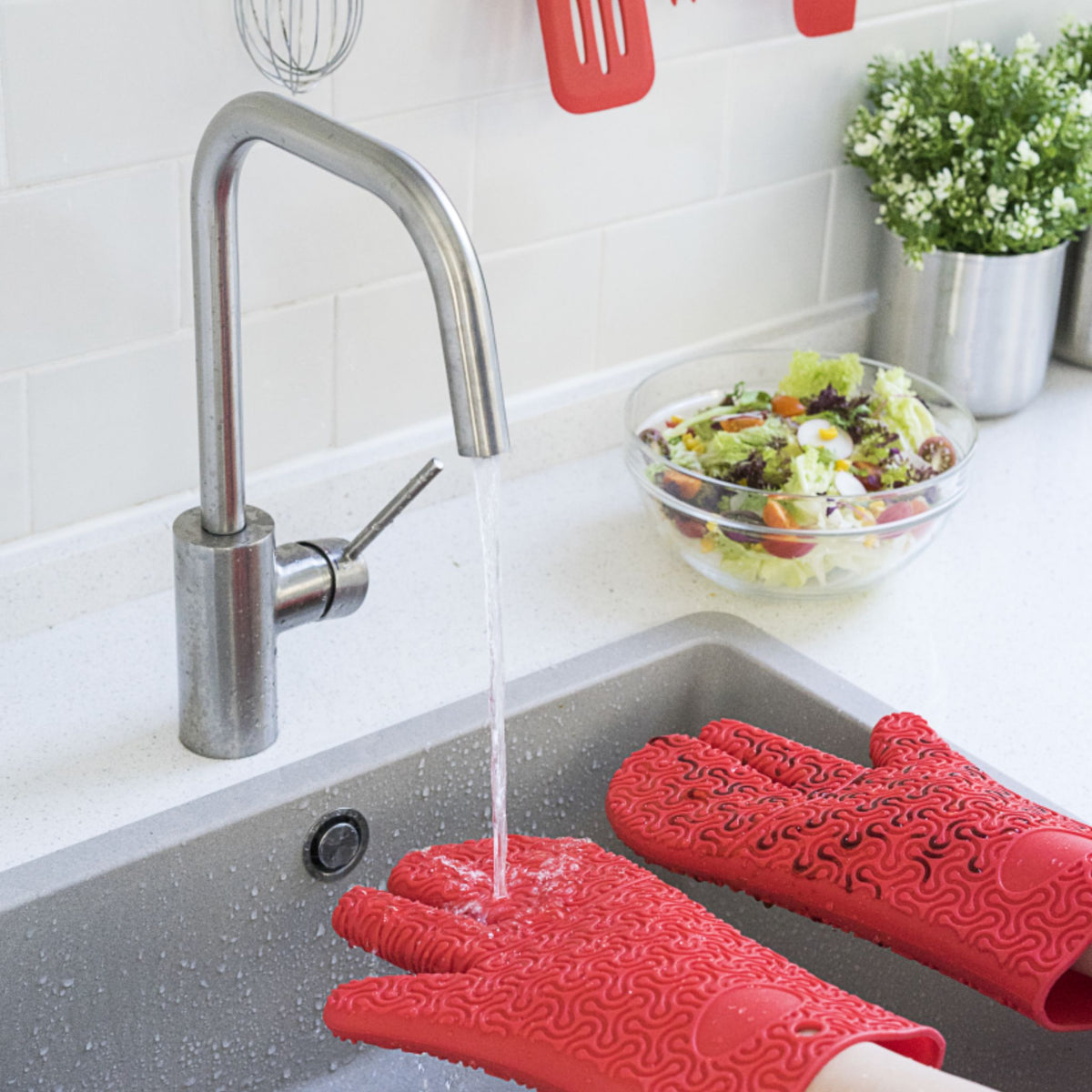 Rote Ofenhandschuhe mit Muster auf der Arbeitsplatte neben Spüle mit Wasserhahn und Salatschüssel.