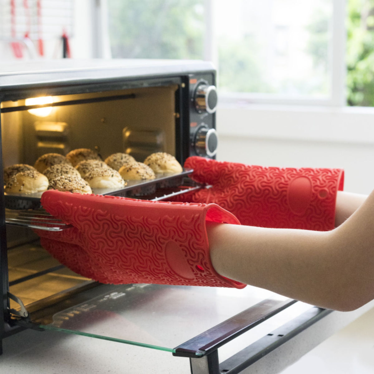 Rote Ofenhandschuhe mit floralem Muster halten Backblech mit Brötchen aus Ofen.