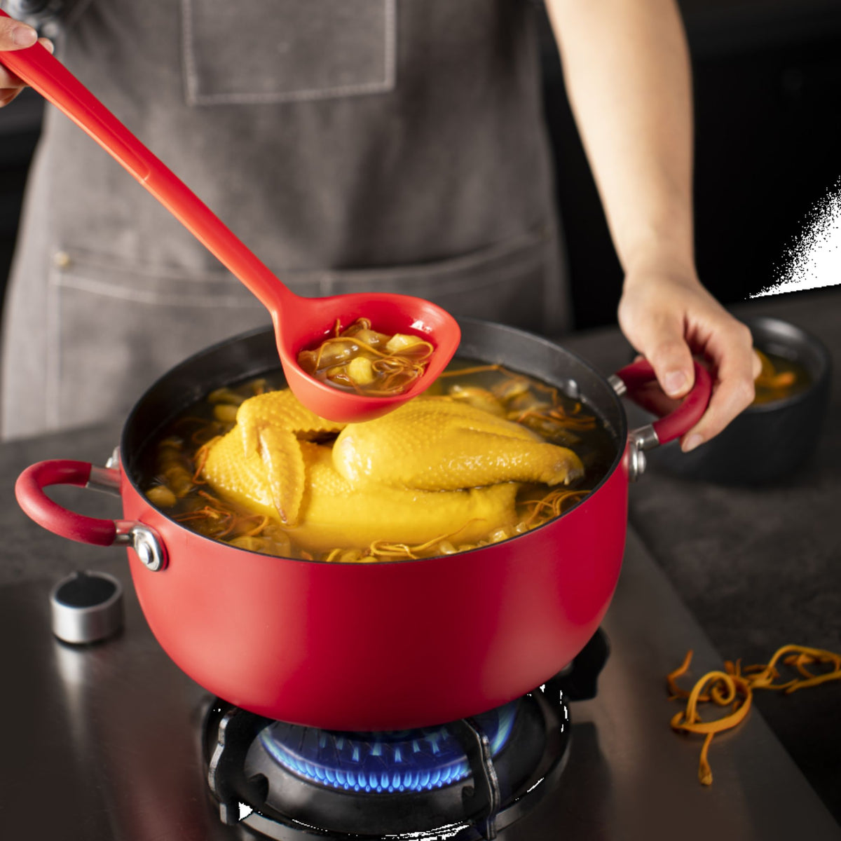 Rote Schöpfkelle mit langem Griff in rotem Topf auf Herd beim Kochen.