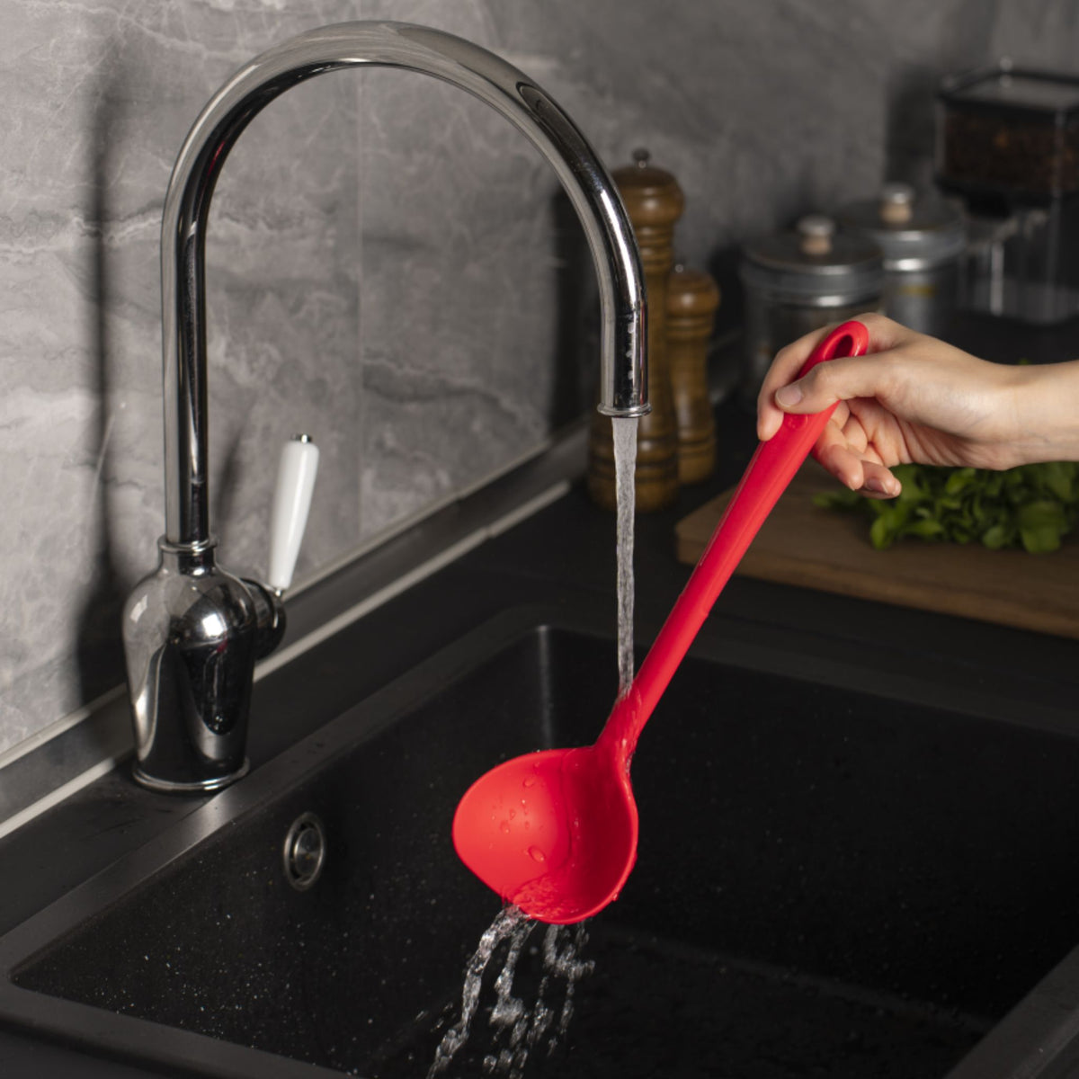 Rote Schöpfkelle aus Kunststoff mit langem Stiel über schwarzer Spüle unter laufendem Wasserhahn.
