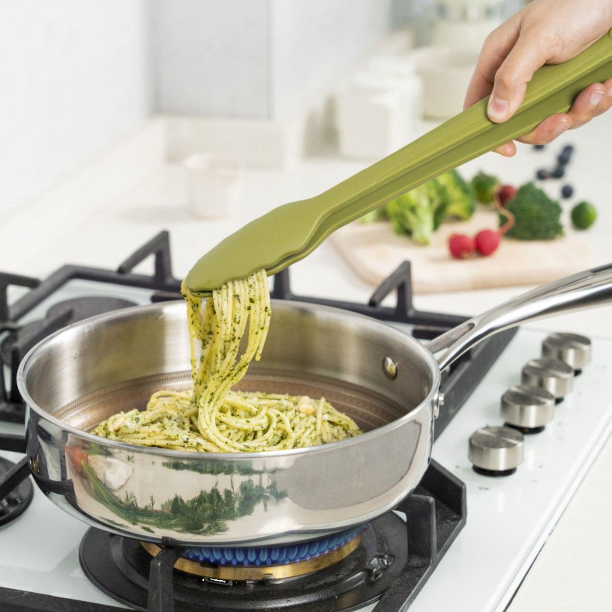 Anthrazitfarbene Grillzange hebt Spaghetti aus Edelstahlpfanne auf Gasherd.