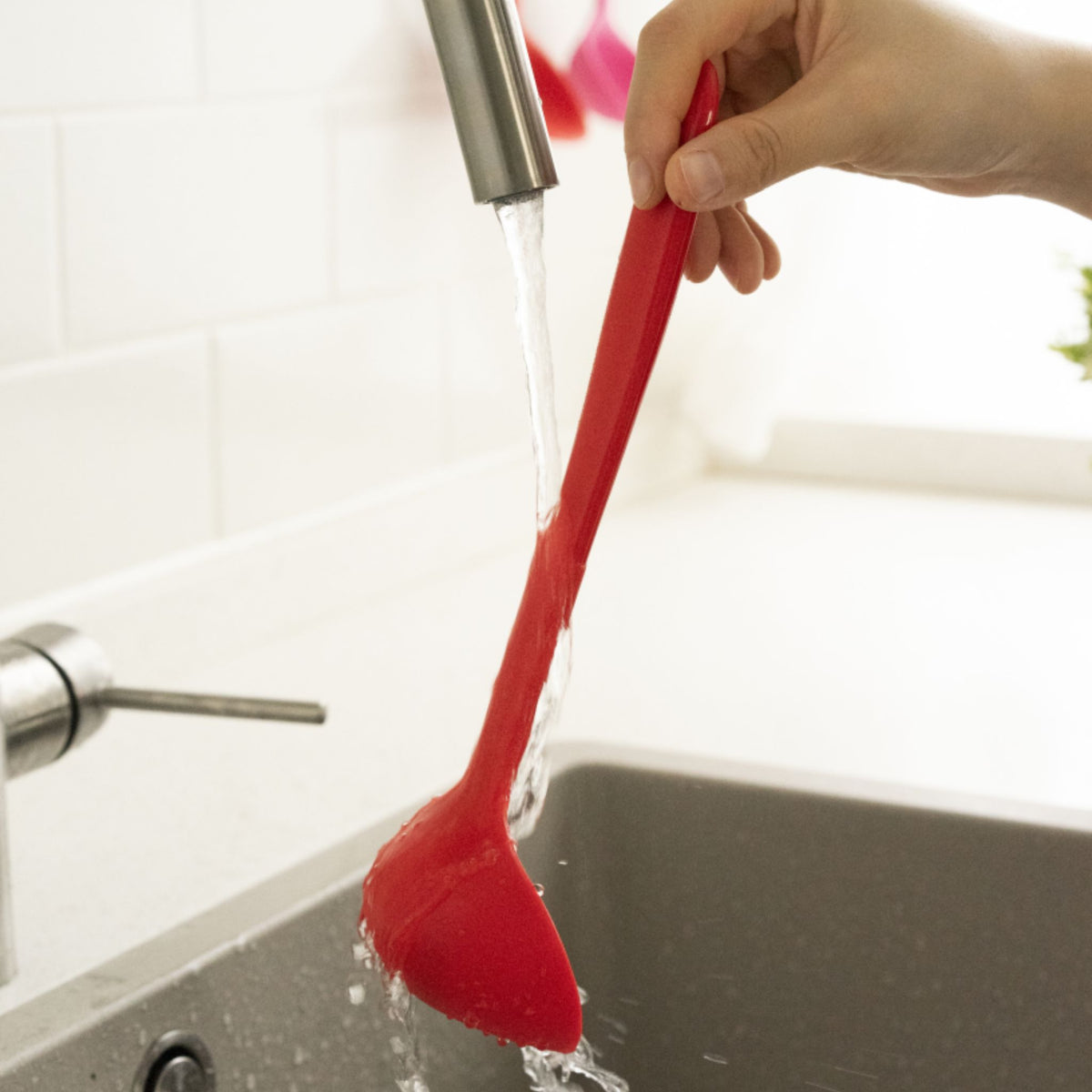 Rote Silikonschöpfkelle mit langem Griff, gehalten über Spüle mit Wasserstrahl.