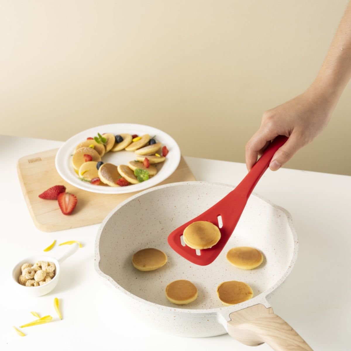 Roter Pfannenwender hebt kleine Pfannkuchen aus weißer Pfanne mit Holzgriff.