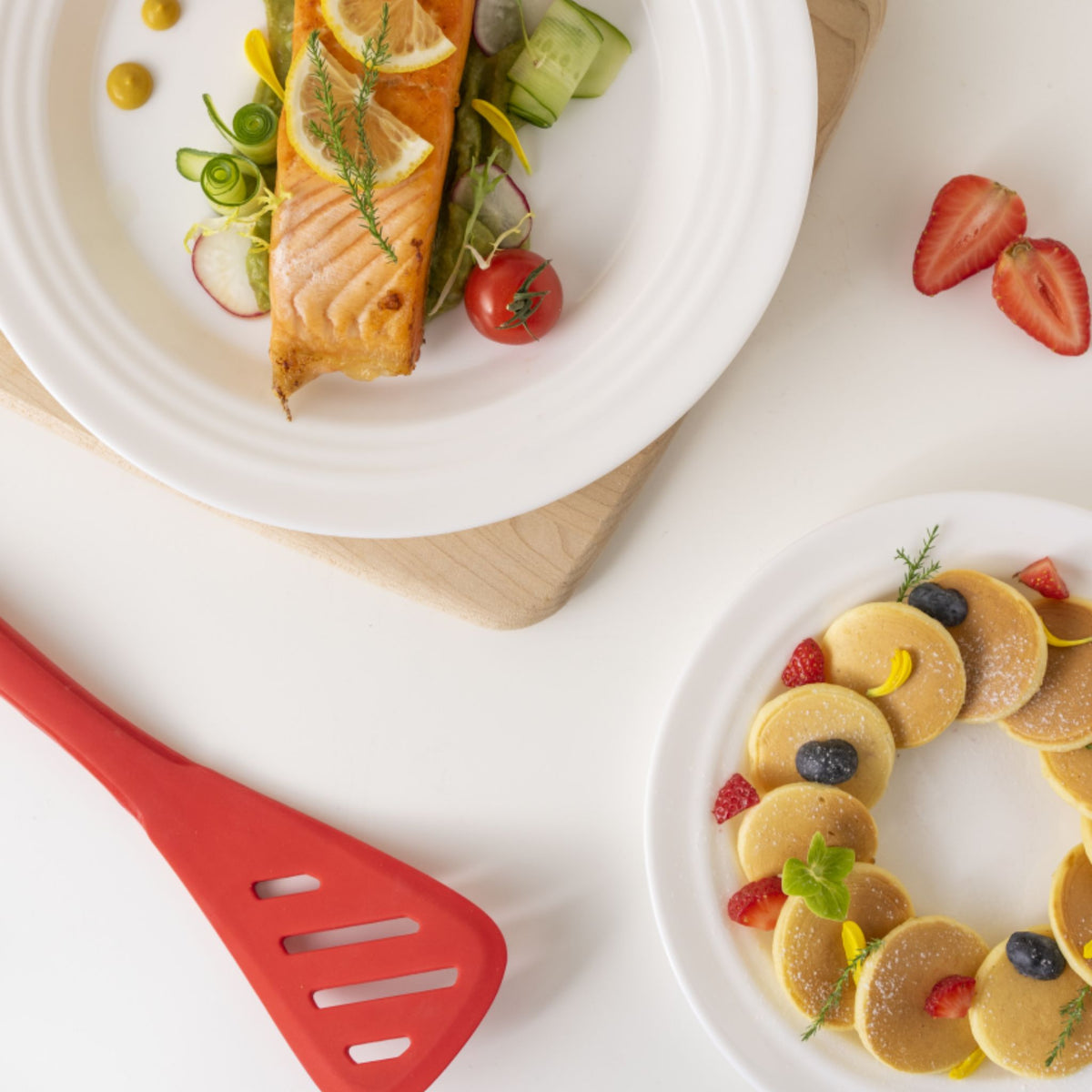 Roter Pfannenwender aus Silikon neben Teller mit Lachs und Beilagen auf weißem Untergrund.