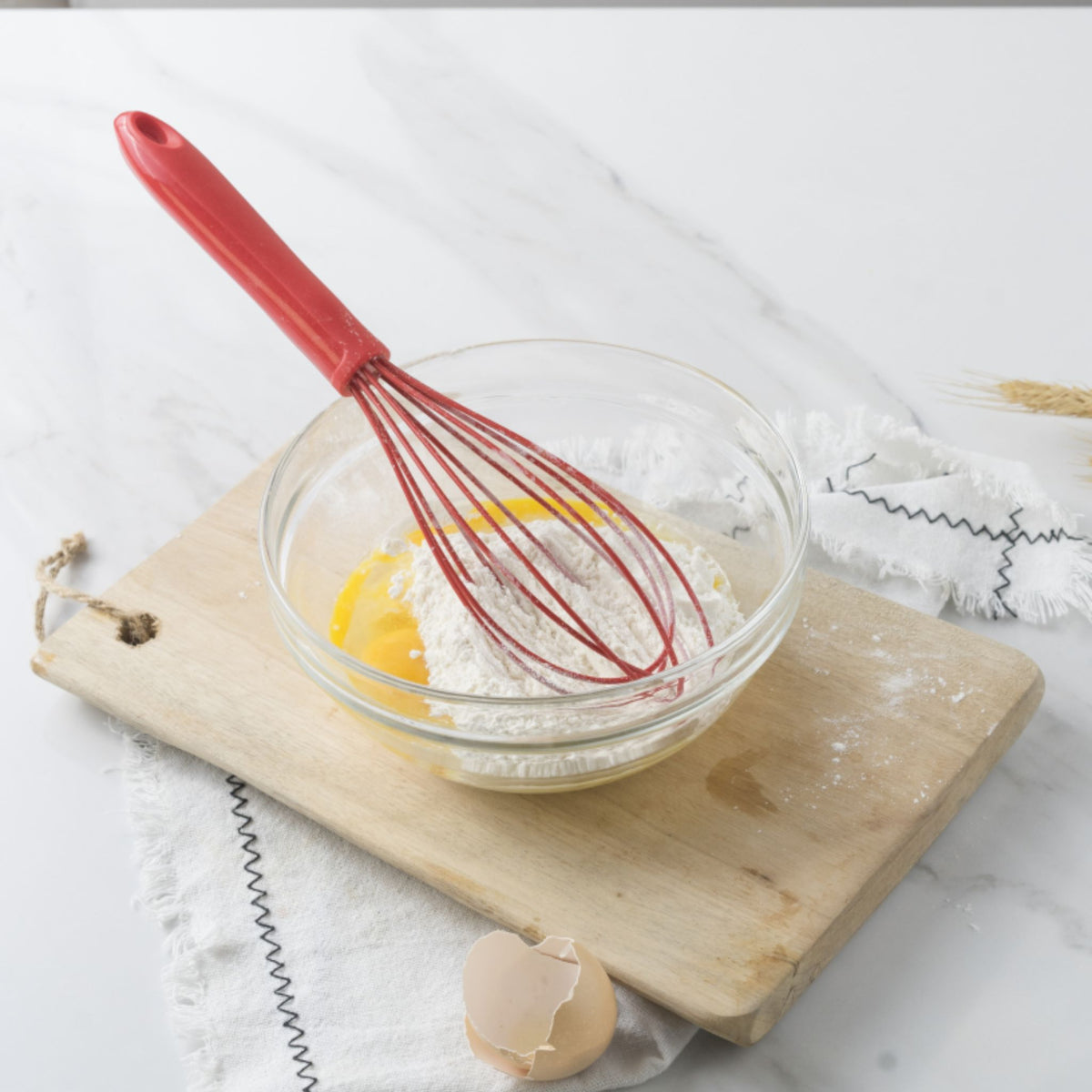 Schneebesen mit rotem Griff rührt Teig in durchsichtiger Glasschüssel auf Holzbrett.