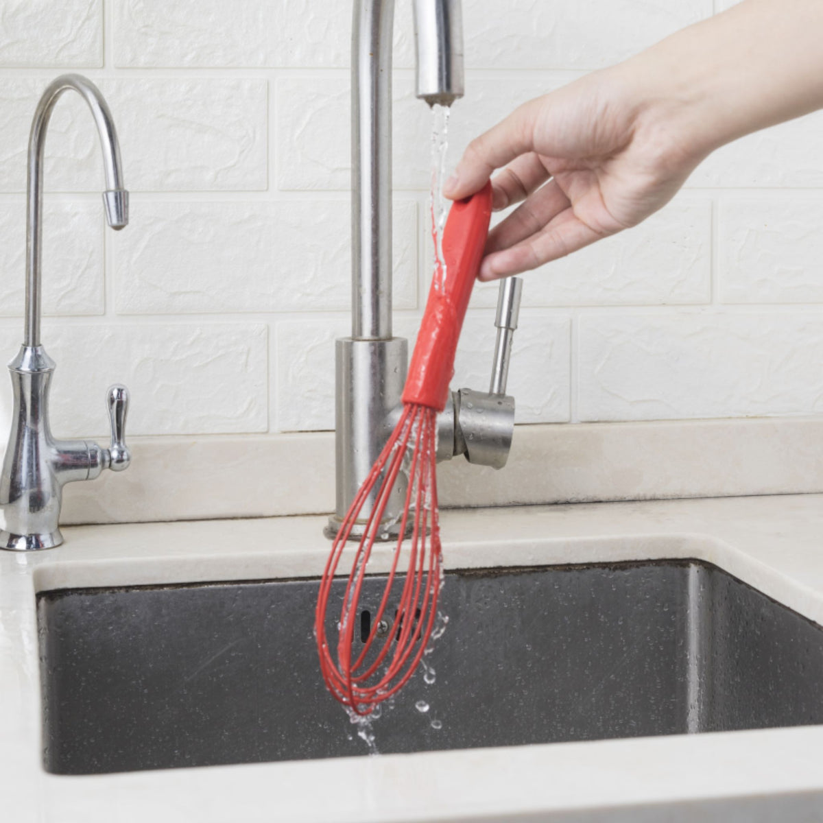 Roter Schneebesen mit ergonomischem Griff wird über Spüle mit Wasser abgespült.