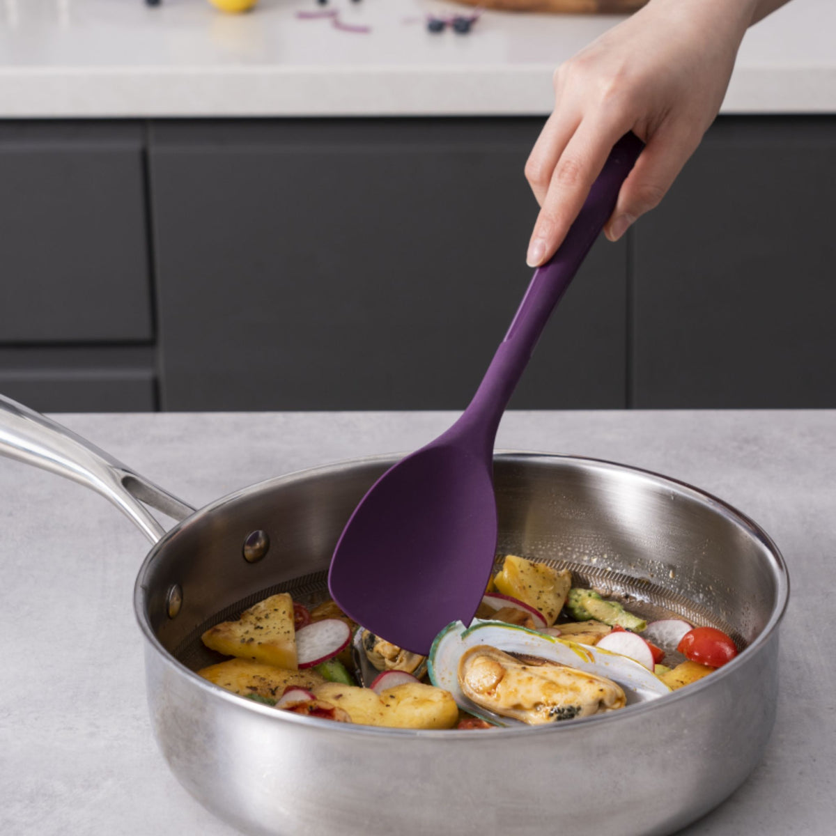 Lila Flexlöffel mit flexiblem Kopf rührt Gemüse in Edelstahlpfanne.