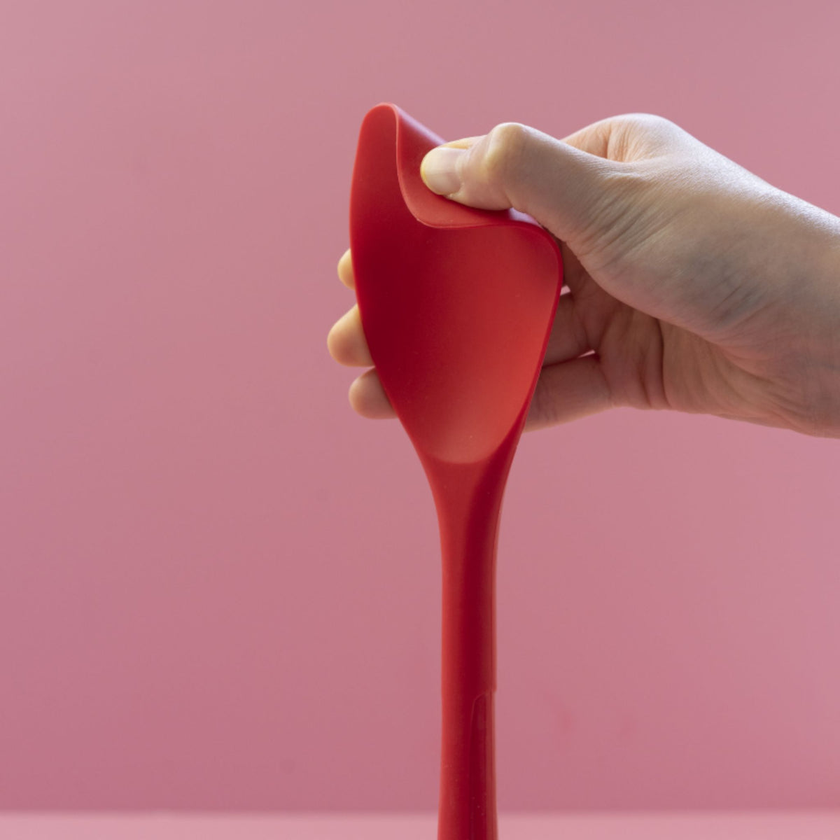 Flexlöffel in kräftigem Rot aus flexiblem Material, gehalten von rechter Hand vor rosa Hintergrund.