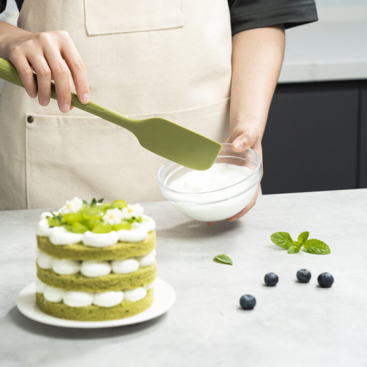 Grüner Teigschaber aus Silikon, gehalten über einer glasschüssel mit weißer creme, daneben ein mehrschichtiger grüner kuchen .