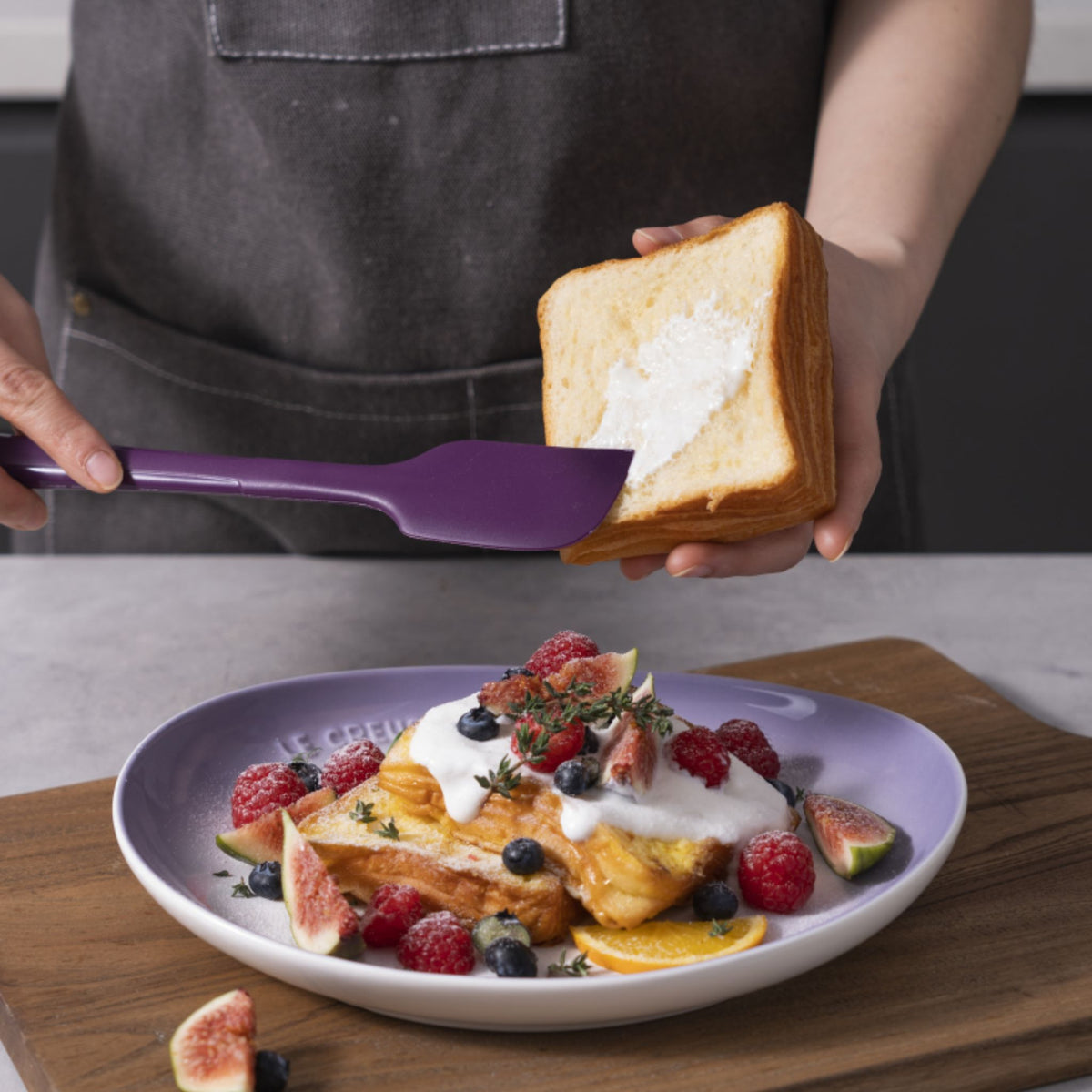 Lila Teigschaber verstreicht Frischkäse auf einer Scheibe Toastbrot.