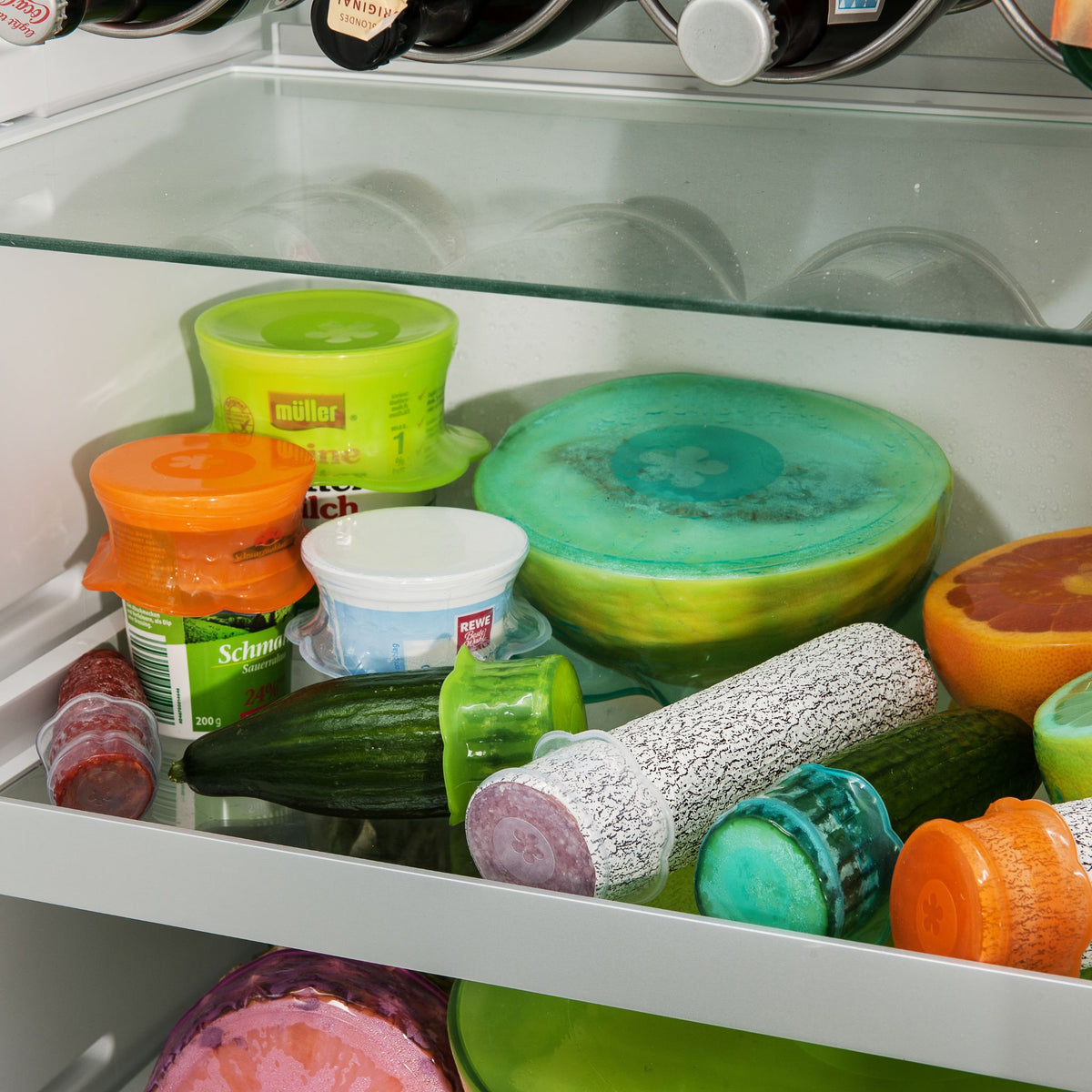 Verschiedene bunte Stretch-Deckel in Orange, Limette, Türkis und Eisblau auf Schüsseln und Lebensmitteln im Kühlschrank.