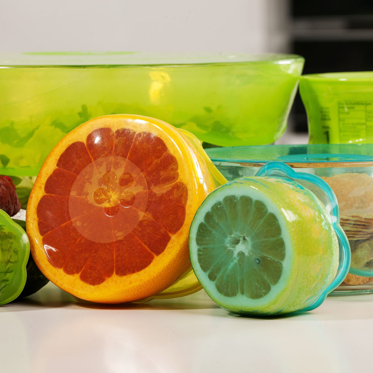 Runde Deckelverschluss-Kappen in Orange und Türkis mit Fruchtmotiv vor grünen Schalen aus Glas.