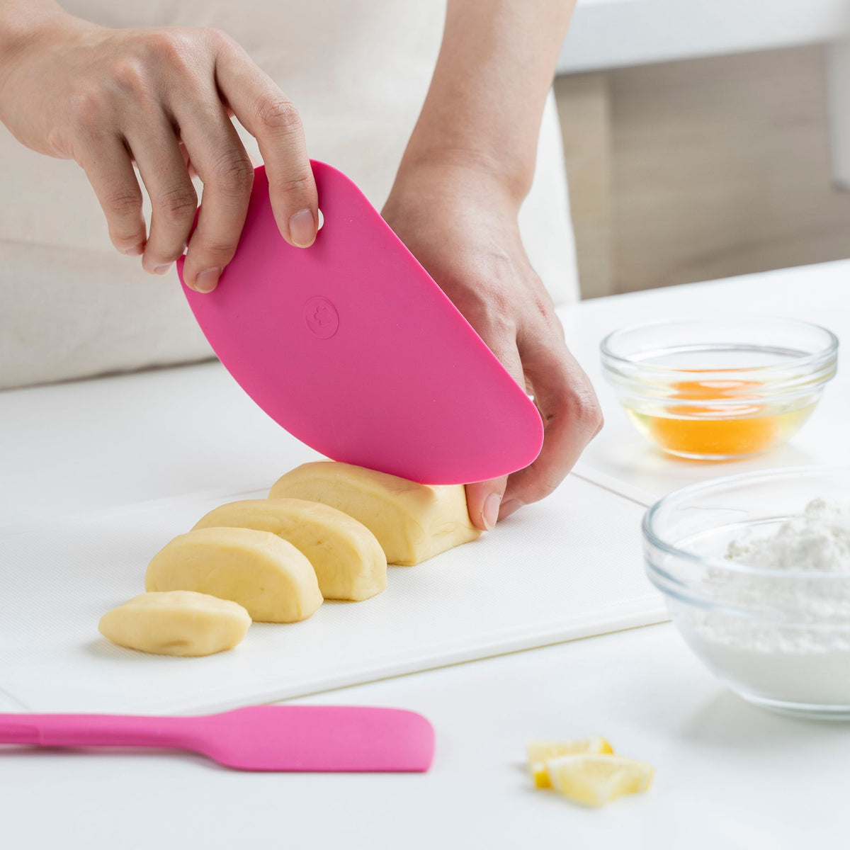 Hand hält pinke Teigkarte, teilt Teigstücke auf hellem Untergrund, Küchenutensilien im Hintergrund.