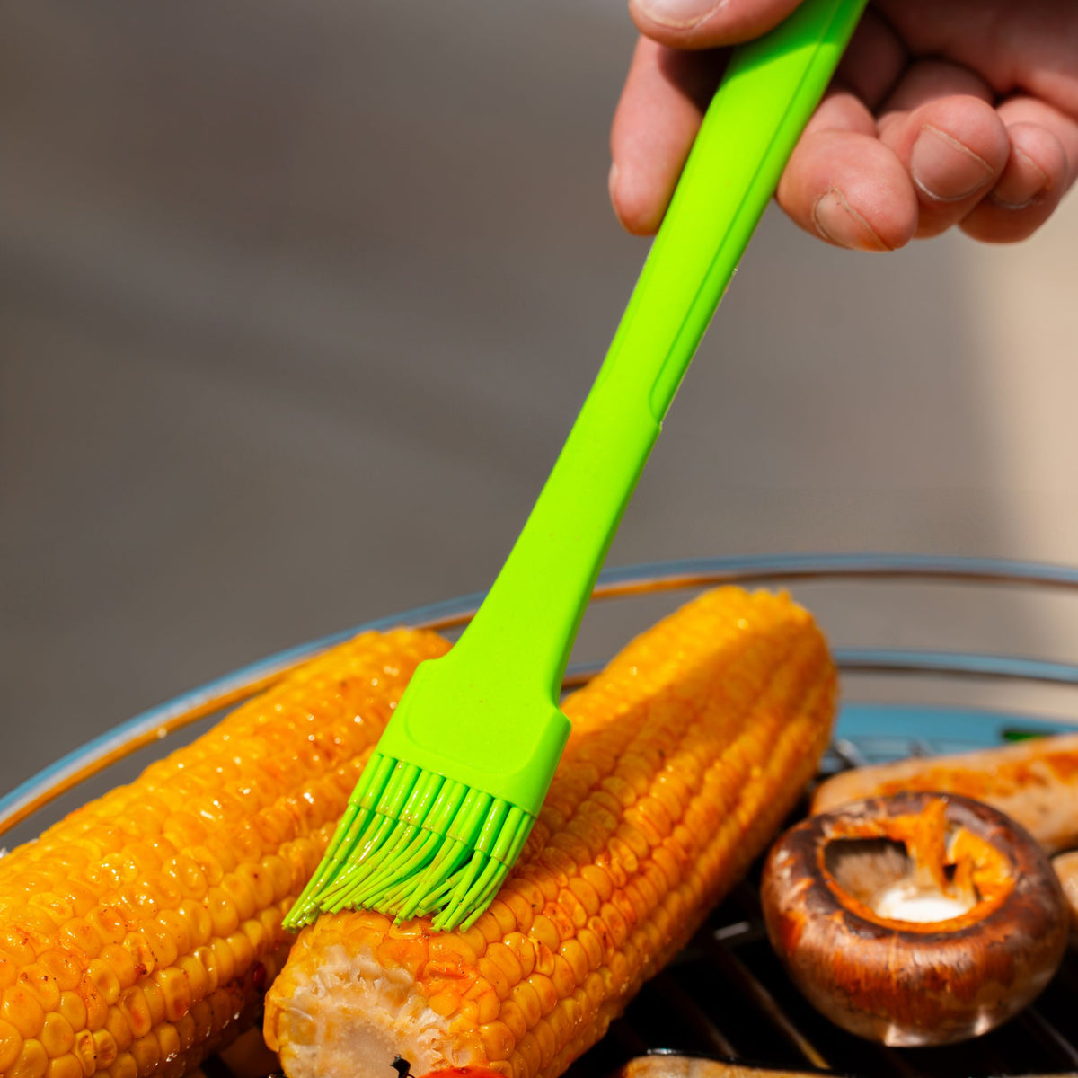 Grüner Backpinsel mit Silikonborsten bepinselt gegrillte Maiskolben auf Grillrost.